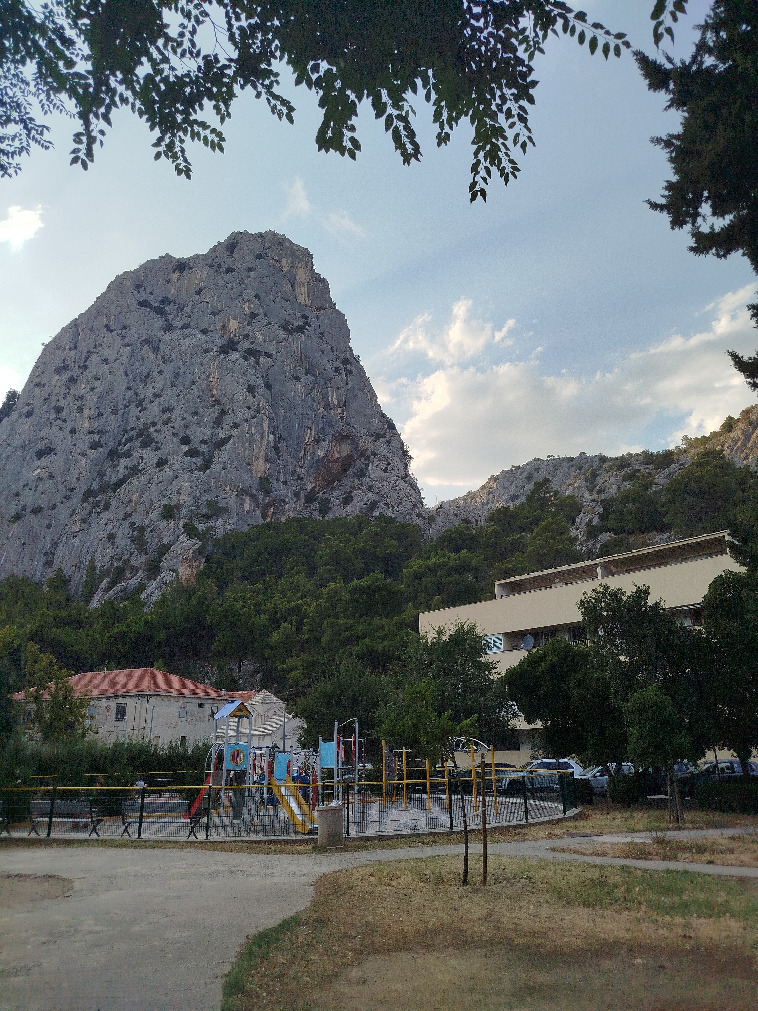 The image depicts a view of the city of Omiš, Croatia, with a prominent rocky mountain dominating the background. In the foreground, there is a children's playground with colorful equipment, including slides and climbing structures, surrounded by a black fence. Benches are visible on the side of the playground. Adjacent to the playground, to the left, are buildings with red tile roofs. To the right, a modern, light-colored apartment building with balconies and satellite dishes is situated. The area around the playground and buildings is a mix of paved paths and dry, grassy areas. Trees and dense green foliage are scattered throughout the scene, with some branches from an overhead tree framing the top of the image. The sky is partly cloudy. No people are clearly visible. The scene appears to be an outdoor recreational area within a residential or semi-residential part of Omiš, with the imposing natural landscape as a backdrop.
