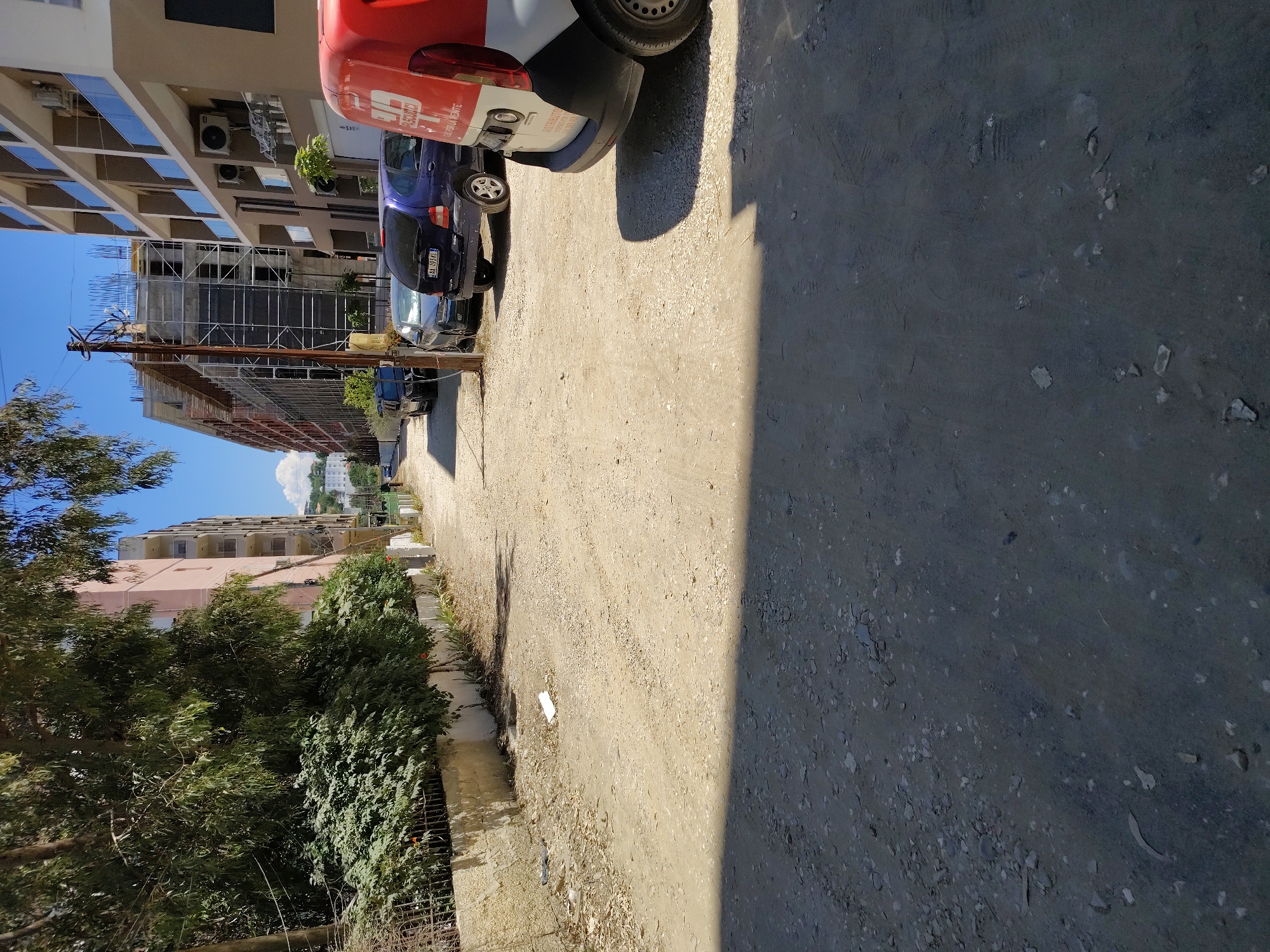 The image depicts a street scene in Kryemëdhenj, Albania, on a sunny day. The street is unpaved, composed of gravel and dirt, with some debris scattered along the sides. On the left, a row of lush green trees and dense bushes lines a low white wall. Beyond the foliage, a light pink building with multiple balconies and windows is visible. To the right, a modern building with a beige facade and glass balconies stands prominently. In front of these buildings, several cars are parked along the street, including a dark blue minivan, a silver sedan, and a purple hatchback with a license plate that reads "AA 959KI". A red and white delivery van is partially visible in the foreground on the right. In the background, a multi-story building under construction, with scaffolding and exposed rebar, signifies ongoing development in the area. The sky is clear and blue with a few scattered clouds.