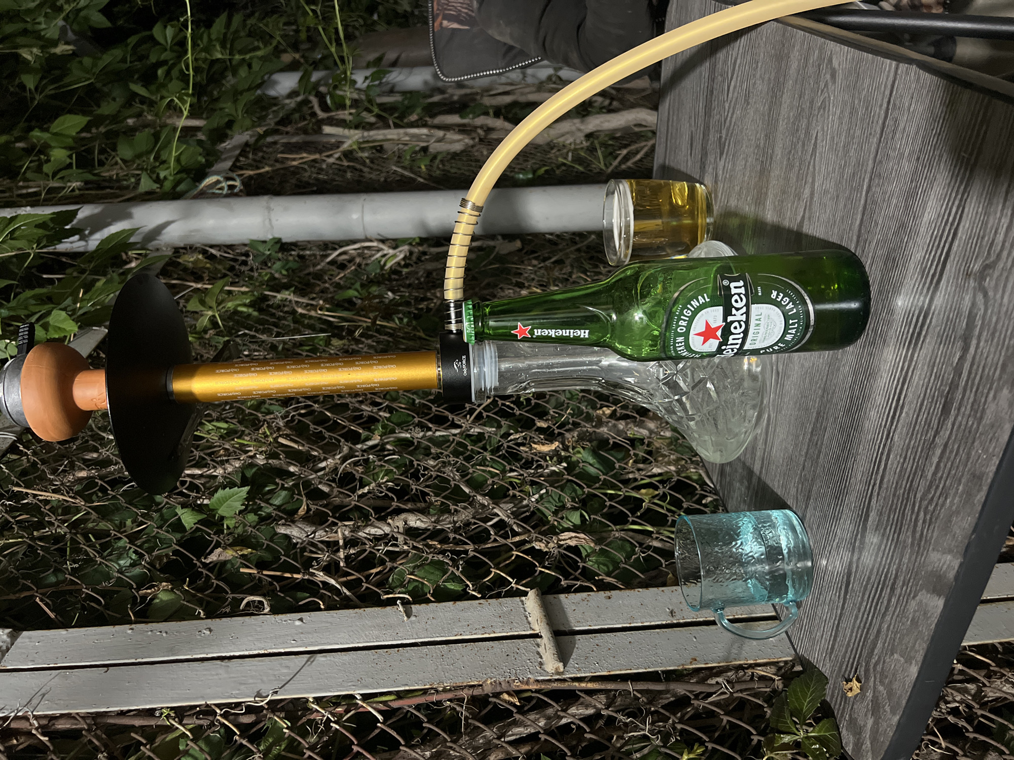 The image depicts an outdoor setting at night, possibly a backyard or patio. A hookah is set up, with its golden stem and black bowl visible. A bottle of Heineken beer stands upright next to a glass containing a yellow liquid, presumably beer. A clear glass mug filled with water is also on the table. The table itself has a weathered wood grain finish. In the background, a chain-link fence is covered with dense green foliage, and a grey pole rises vertically. The overall atmosphere suggests a casual, relaxed evening. There are no identifiable individuals present in the scene. The location is specified as Baikonur, Kazakhstan.