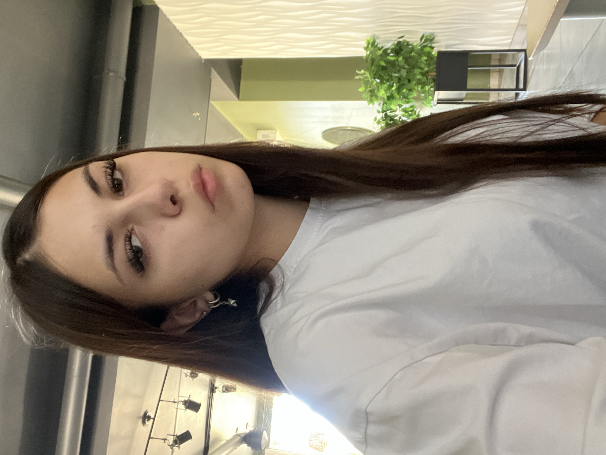 A young woman with long, dark brown hair is captured in a selfie-style, close-up shot. She is wearing a plain white t-shirt and silver hoop earrings, one of which has a star-shaped charm. Her gaze is directed slightly away from the camera, with a neutral expression. The background suggests an indoor, possibly public space with a modern aesthetic. Visible elements include a ceiling with exposed industrial lighting fixtures, a textured white wall with a wave-like pattern, a tall green plant in a black planter, and a hint of a wooden surface, potentially a table or counter. The lighting appears to be a mix of natural and artificial sources, casting subtle shadows. The overall impression is casual and unposed. The location of Красные Баки, Russia is not ascertainable from the visual information provided.