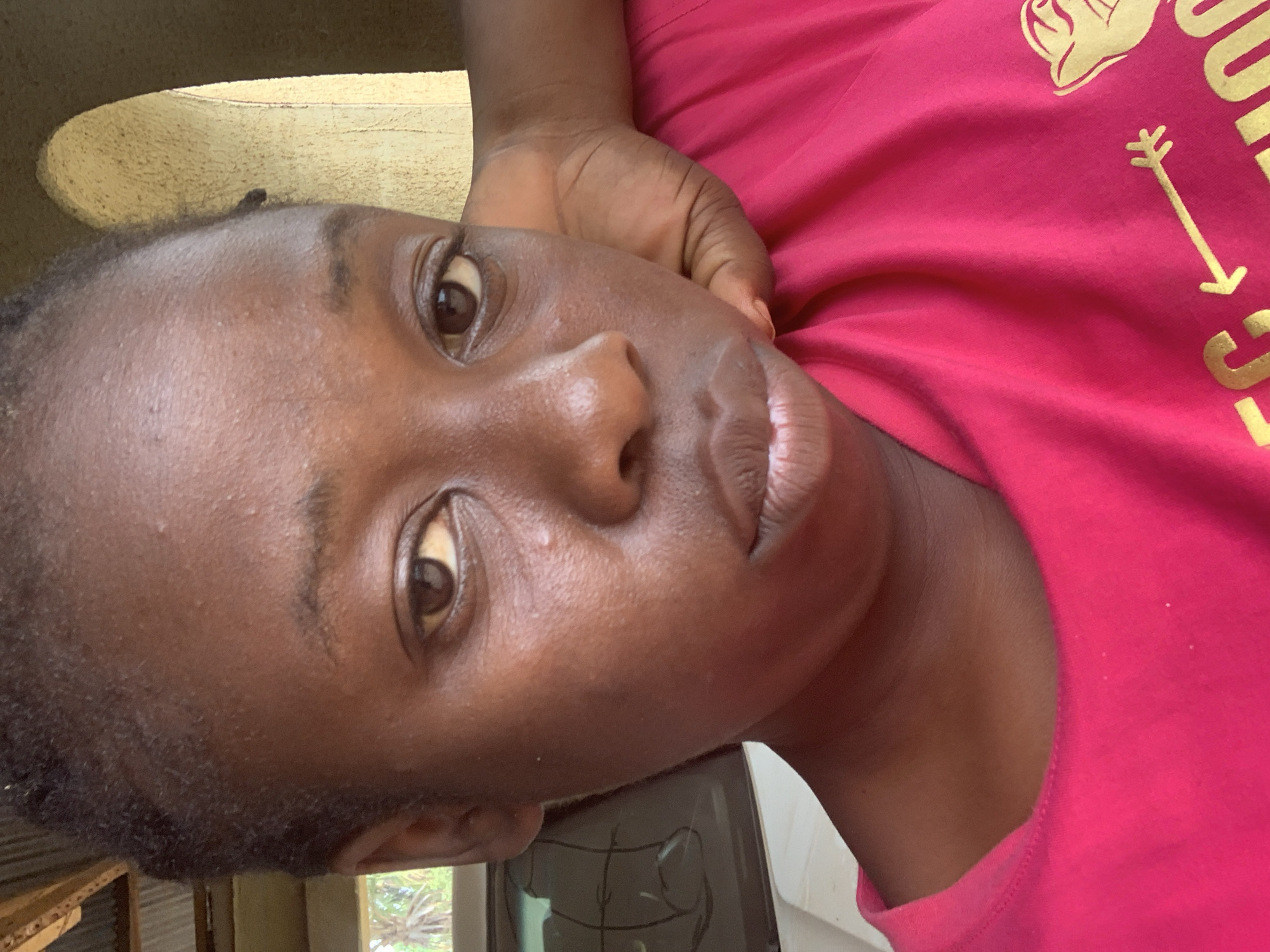 A close-up portrait features a young individual with dark skin, directly facing the camera, within the city of Akwanga, Nigeria. The subject wears a bright fuchsia-colored t-shirt with a rounded neckline, displaying a partial yellow graphic design on the lower right, including an arrow pointing left and indistinct text or imagery. Their left hand is visible, bent at the wrist, positioned behind their head or neck. The background on the left reveals corrugated metal roofing, a segment of a white vehicle with a dark window showing scribbled marks or reflections, and green palm-like foliage. On the right, a light yellowish, textured arched opening or doorway is visible, indicative of a building structure. Natural daylight illuminates the scene.