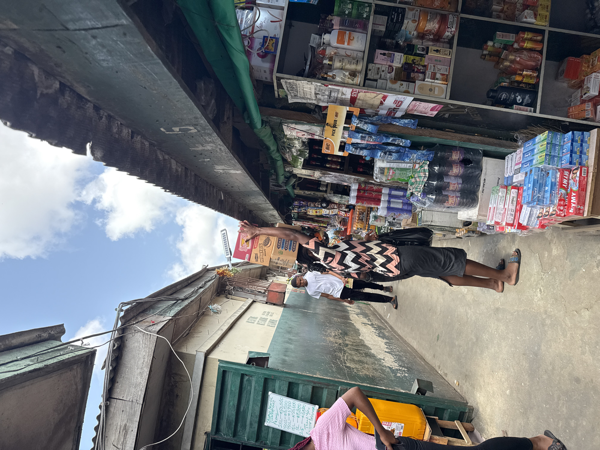 The image captures a vibrant, narrow street market scene in Lekki, Nigeria, during the daytime under a bright, cloudy sky. The perspective looks down a pathway flanked by commercial activity.
On the right side of the street, a bustling shop or stall is crammed with diverse merchandise. Shelves are densely packed with products including cosmetics (like Nivea, Dr. Rashel, Visita Plus), toiletries (Oral B, Pepsodent, MYMY), and packaged goods. A prominent "Bic" display invites customers to "Pick Your Bic," and cases of "Oral B" products, as well as bottles labeled "drink" with "40% Alc Vol.," are visible.
Several individuals are present on the concrete pathway. In the mid-ground, a person wearing a black and white patterned top and dark shorts walks away from the camera, skillfully balancing a cardboard box on their head. Further down the street, another person in a white t-shirt and black pants can be seen. In the foreground, to the left, a person in a pink top and black pants stands next to a large yellow container. Adjacent to them is a white signboard detailing prices for "ORGANIC PALM OIL CONTENT ONLY PROMO," with quantities and prices listed in Naira, such as "25L - N52.500" and "5L - N10,800." The sign concludes with "Thank you f your Patronage."
The left side of the street features weathered buildings with corrugated metal roofs and exposed electrical wiring, some covered by green tarpaulins providing shade. The scene depicts a typical bustling market environment, indicative of daily commerce in the area.