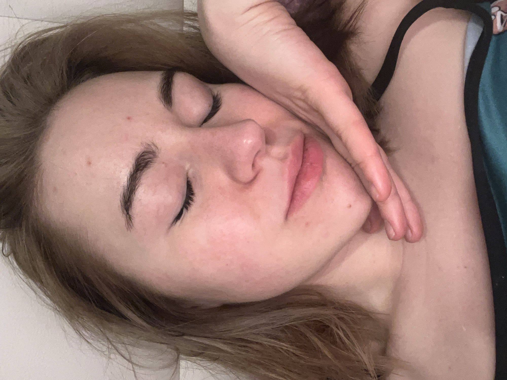 A close-up, eye-level photograph depicts an individual with light brown hair and fair skin, eyes closed, resting in an indoor setting. The location is stated as Boreti, Montenegro, though not visually ascertainable from the image. The subject's head is tilted slightly to the right (viewer's left), supported by their right hand, with fingers curled under the jawline and the palm against the side of the face. The individual's face presents light pink, closed lips, dark, full eyebrows, and visible black eyeliner on the upper eyelids. Small red blemishes are present on the forehead and chin, and a small mole is visible on the right cheek. A dark strap from a garment and a teal/dark green fabric are visible on the lower right portion of the frame, near the shoulder. The background consists of a plain, light-colored surface.