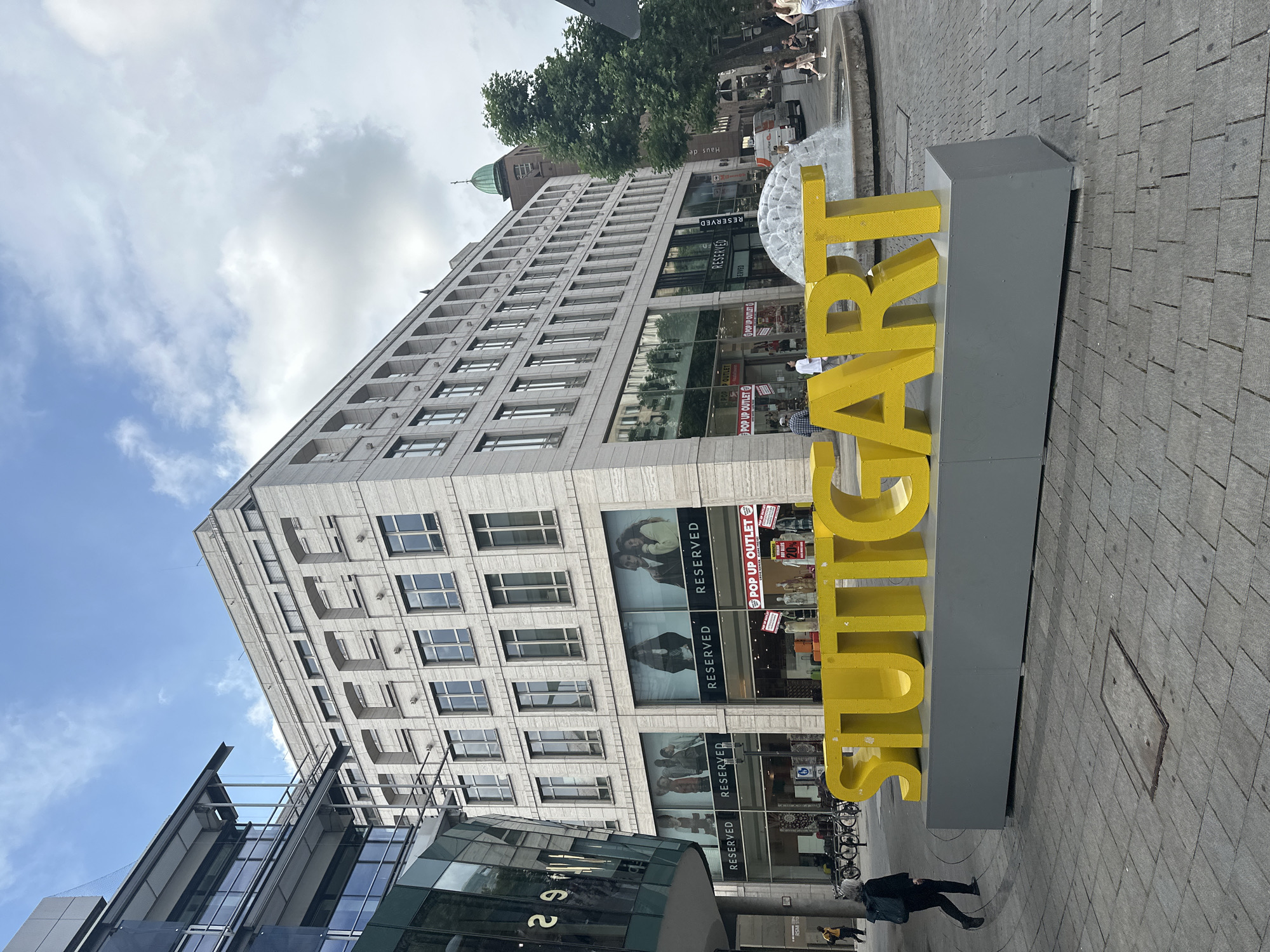 A low-angle, full outdoor shot shows a large, bright yellow "STUTTGART" sign in the foreground, resting on a gray rectangular base. The sign is positioned on a paved pedestrian area, which appears to be a city square. Behind the sign, a modern building with a beige facade and numerous windows dominates the mid-ground. Below the windows on the ground floor, large storefronts are visible, including "RESERVED" and "POP UP OUTLET" signs. Several bicycles are parked on the left side of the image. Further in the background, a fountain with water spraying upwards is partially visible. To the right of the fountain, trees and a building with a green dome can be seen. The sky above is partly cloudy. A few people are visible in the scene, including a person walking away from the camera on the left, and others strolling in the background near the fountain and shops. The overall impression is that of a lively urban environment.