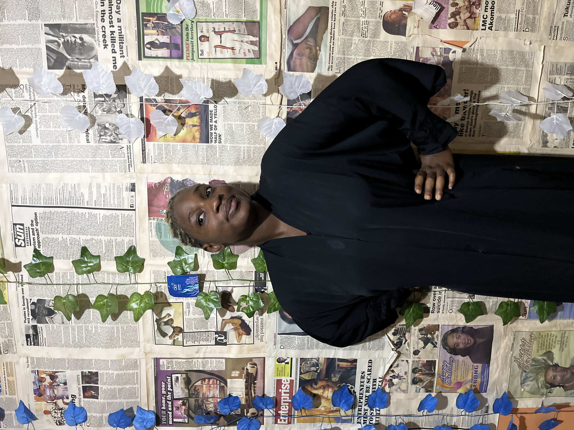 The image depicts a young Black woman standing in front of a wall covered in newspaper clippings. She is wearing a long, flowing black garment and has her left hand on her hip. Her hair is styled in braids, and she is looking slightly off-camera. The newspapers on the wall feature various headlines and images, including portraits of individuals and news-related graphics. There are also artificial green and white leaves attached to the wall, some of which appear to be part of a vine. The lighting suggests an indoor environment. The provided crops show more detail of the newspaper content and the woman's facial features. The location of Rafin Par, Nigeria, is not explicitly visible in the image itself, but the content of the newspapers, such as the Nigerian flag and headlines in English, suggests a Nigerian context.