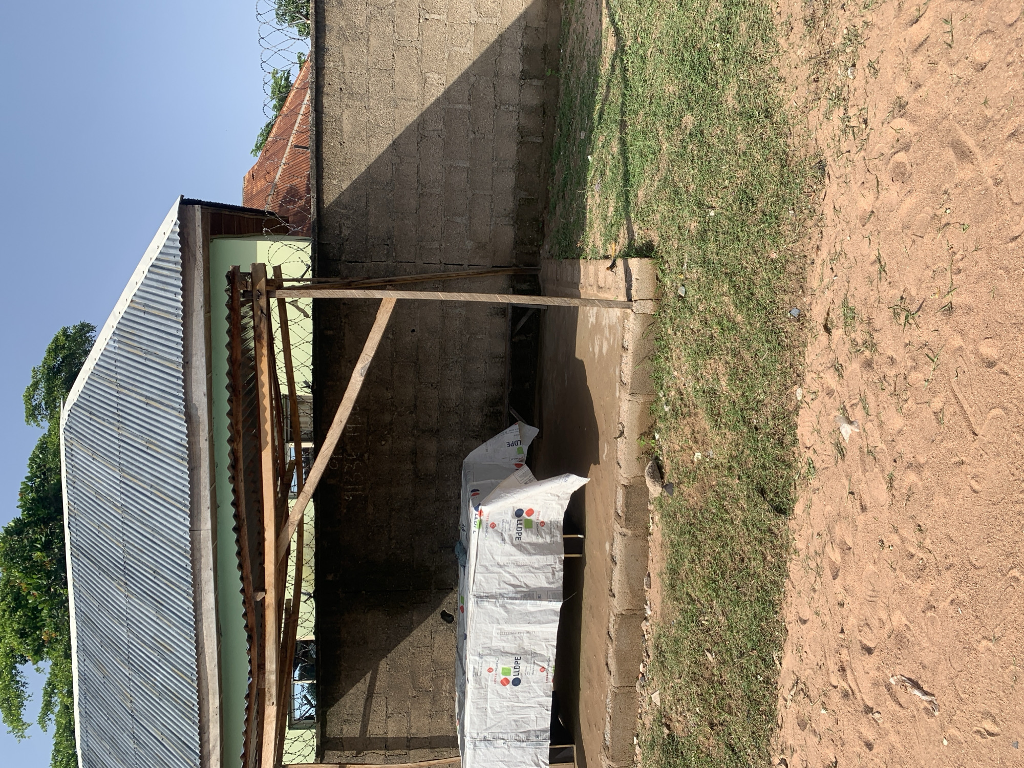 The image depicts an outdoor scene in Jalingo, Nigeria, featuring a covered area with a table underneath. The structure consists of a corrugated metal roof supported by wooden posts, set against a concrete block wall. A table covered with a white tarp, printed with logos and text, is positioned within this covered space. The ground surface around the structure is a mixture of dirt and sparse grass. In the background, the corrugated metal roof of a building is visible, along with a clear blue sky. The scene appears to be situated in an open area, possibly a marketplace or an outdoor gathering space, with elements of both construction and natural environment present.