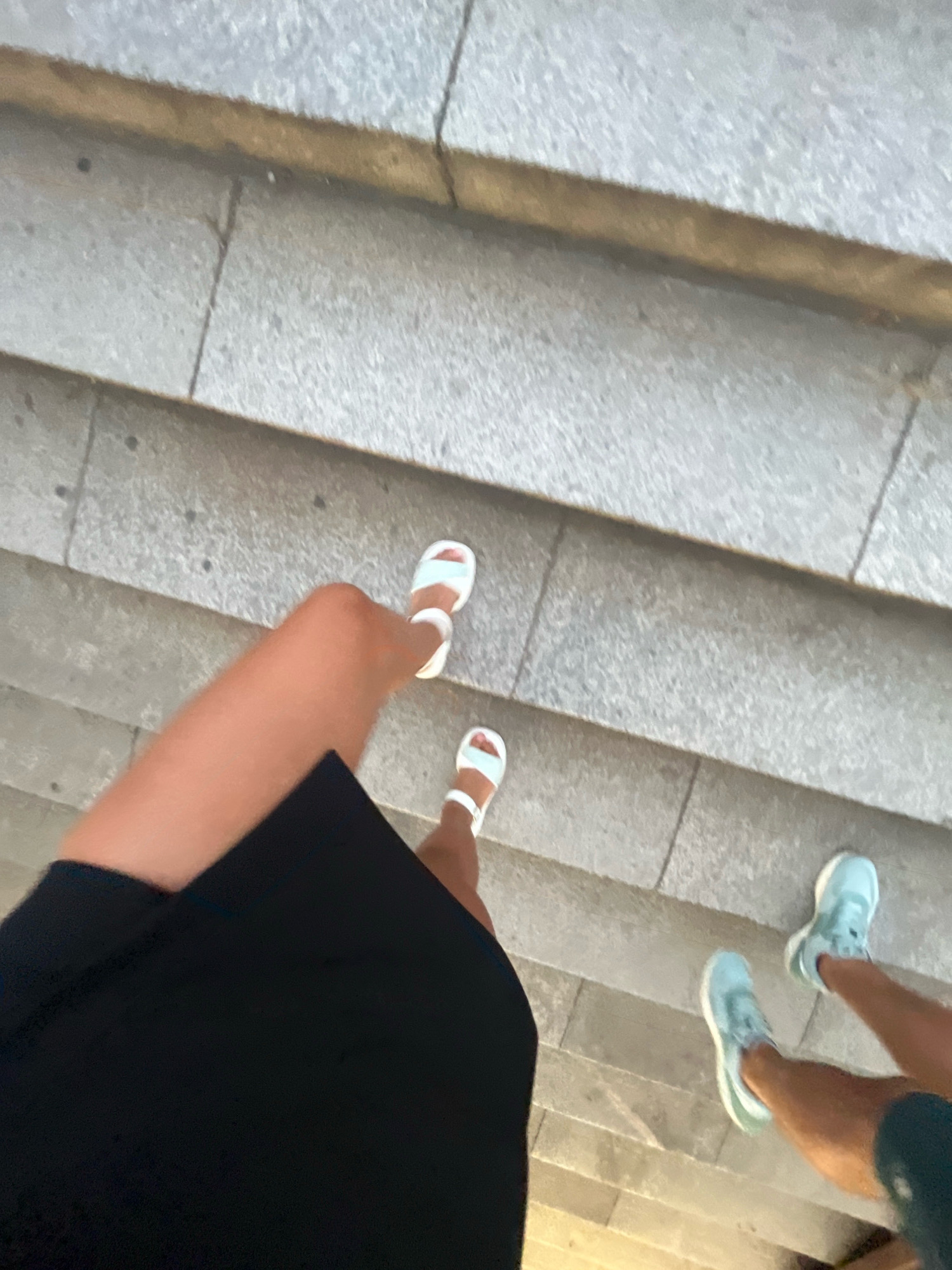 The image shows a first-person perspective looking down at two pairs of legs descending a stone staircase. The person in the foreground is wearing a black skirt and white sandals. To their right, another person is wearing light blue sneakers. The staircase appears to be outdoors, with rough-textured, grey stone steps. The scene could be part of the historic Old Town of Plovdiv, Bulgaria, where many similar stone staircases are found, leading to various landmarks and architectural sites. The lighting suggests it is either early morning or late afternoon.