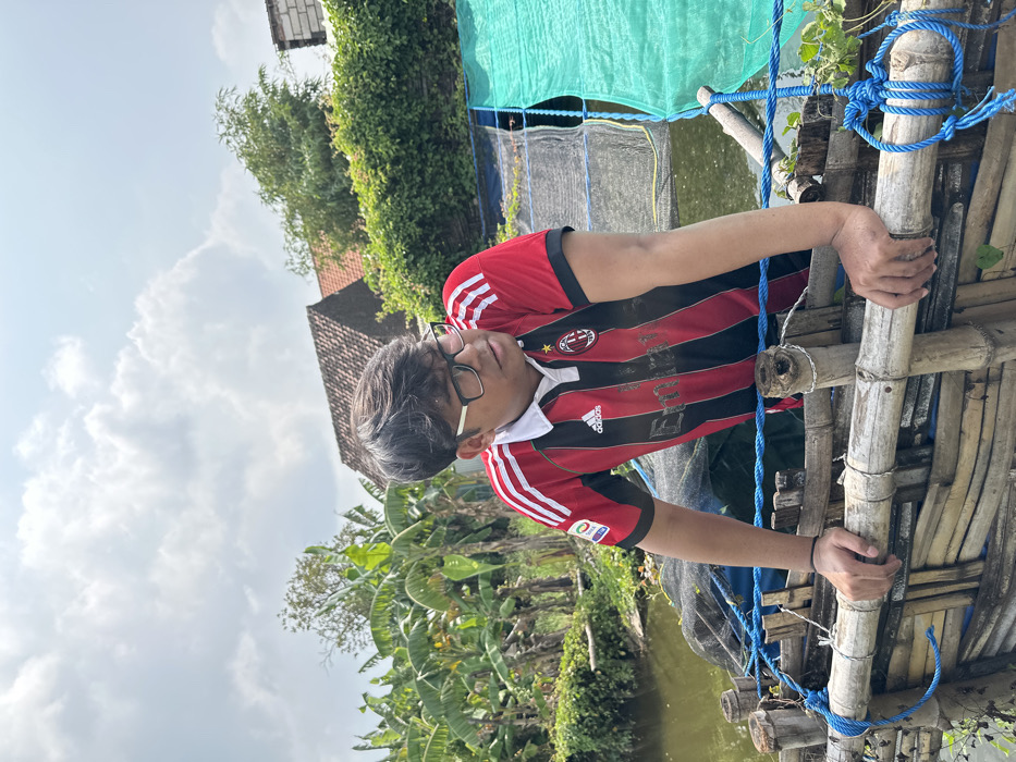 A person wearing glasses and an AC Milan jersey stands on a bamboo structure over a body of water. They are holding onto the structure, which is secured with blue rope. Lush green vegetation is visible in the background, along with a building with a red tiled roof. The sky is cloudy.