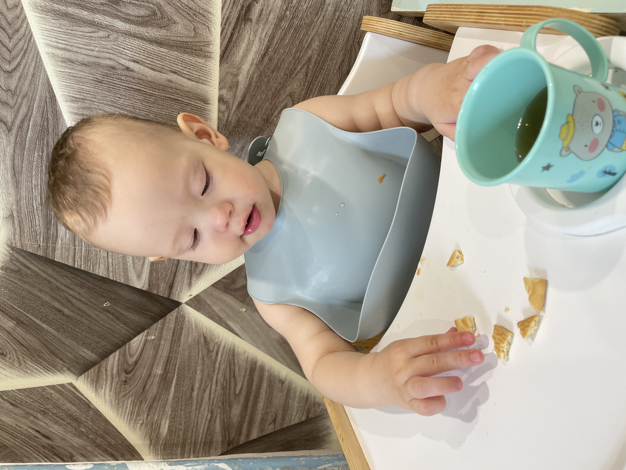 A baby is sitting in a high chair, wearing a gray bib, with small pieces of food scattered on the white tray in front of them. There is a light blue mug with a bear illustration on a white saucer to the right of the baby. The baby's eyes are closed, and they appear to be reaching for a piece of food. The background shows a wall with a patterned wood design. The location is Perm, Russia.