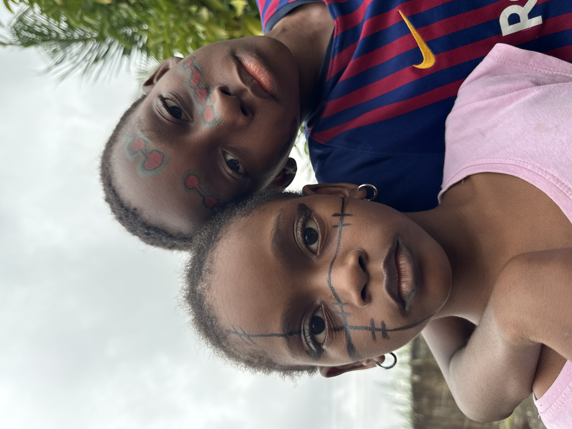 Two young Black individuals, likely children or adolescents, are depicted in a close-up portrait, their faces adorned with face paint. The individual in the foreground, positioned on the left, has dark skin and black hair. Their face features black markings, including a vertical line down the center of their forehead and across the bridge of their nose, with horizontal lines extending from the nose to the cheeks, and additional markings resembling eyelashes and lines under the eyes. They are wearing a silver hoop earring in their left ear and a light pink t-shirt.
The second individual, positioned slightly behind and to the right, also has dark skin and dark, curly hair. Their face paint consists of red and green designs, with circular elements and lines around their eyes and forehead. They are wearing a blue and red striped t-shirt with a yellow Nike logo and what appears to be a "R" on the right side. The two individuals are positioned closely, with the second individual's head leaning towards the first's shoulder. The background is an outdoor setting with green foliage visible, suggesting a natural or semi-natural environment. The sky above is overcast and bright. The overall impression is a posed photograph.