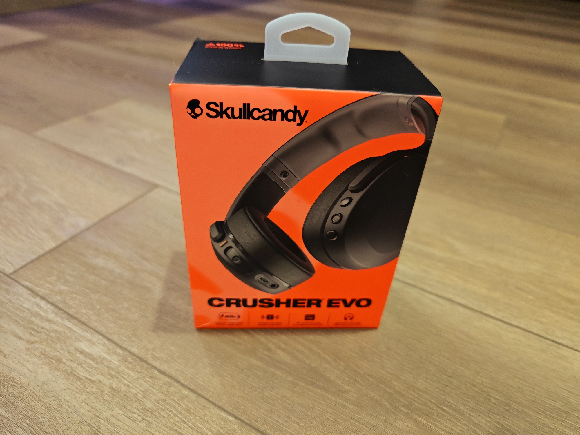 An orange and black Skullcandy Crusher Evo headphones box is placed on a light brown wooden floor. The box has the Skullcandy logo and a picture of the headphones on its front. The top of the box has a white plastic handle. The floor appears to be indoors and shows natural wood grain. The scene is well-lit. There is no information to determine the location in Calgary, Canada.