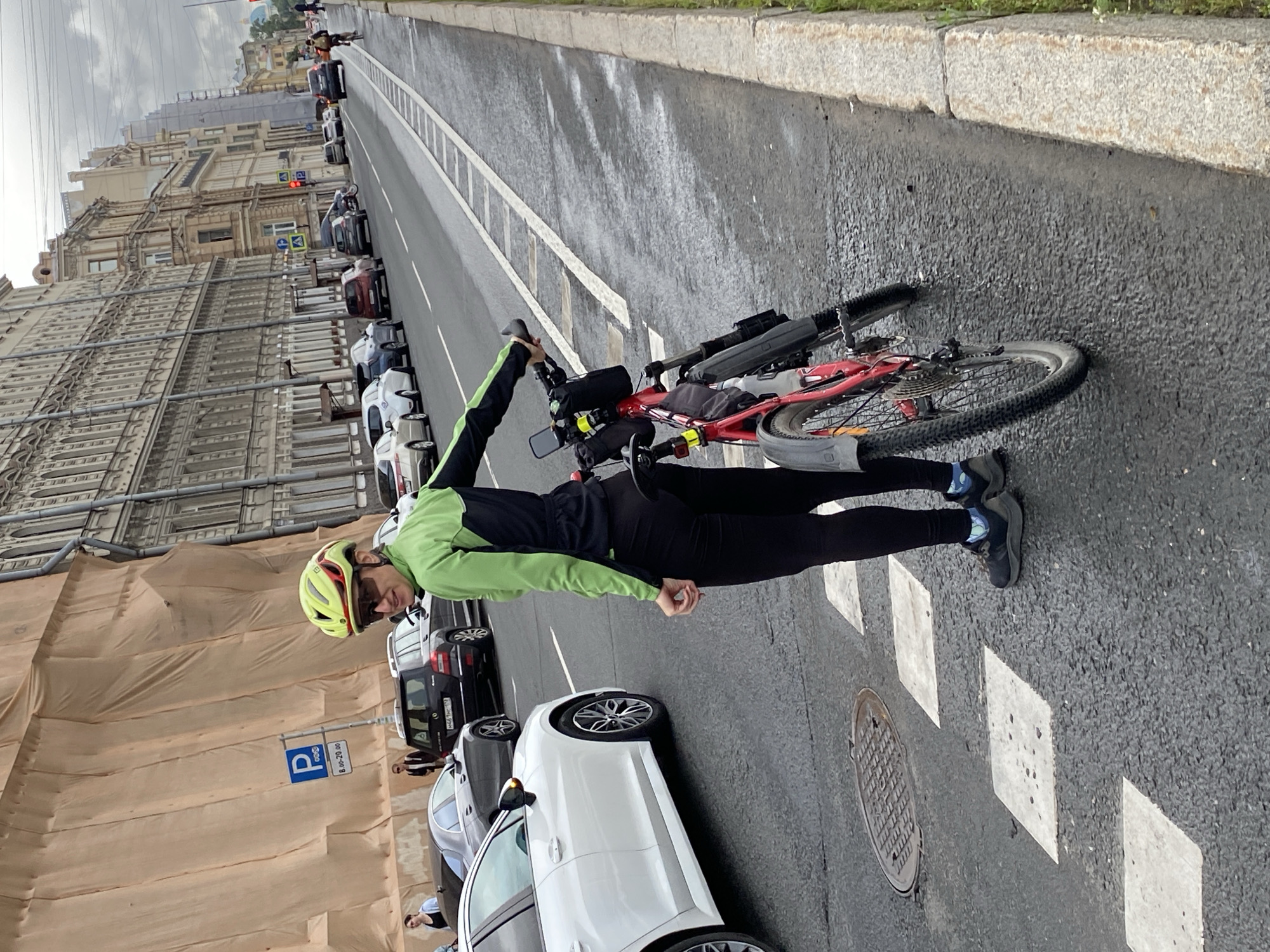 A woman wearing a neon green and black cycling jacket, black leggings, a yellow helmet, and sunglasses stands on a pedestrian crossing on a street in Saint Petersburg, Russia, holding the handlebars of a red bicycle. The bicycle has a black saddle bag and a water bottle cage. The street is wet, indicating recent rain. Buildings line the street, and cars are parked and driving on the road. A large building with scaffolding and a beige tarp is visible on the left side of the image. The sky is overcast.
