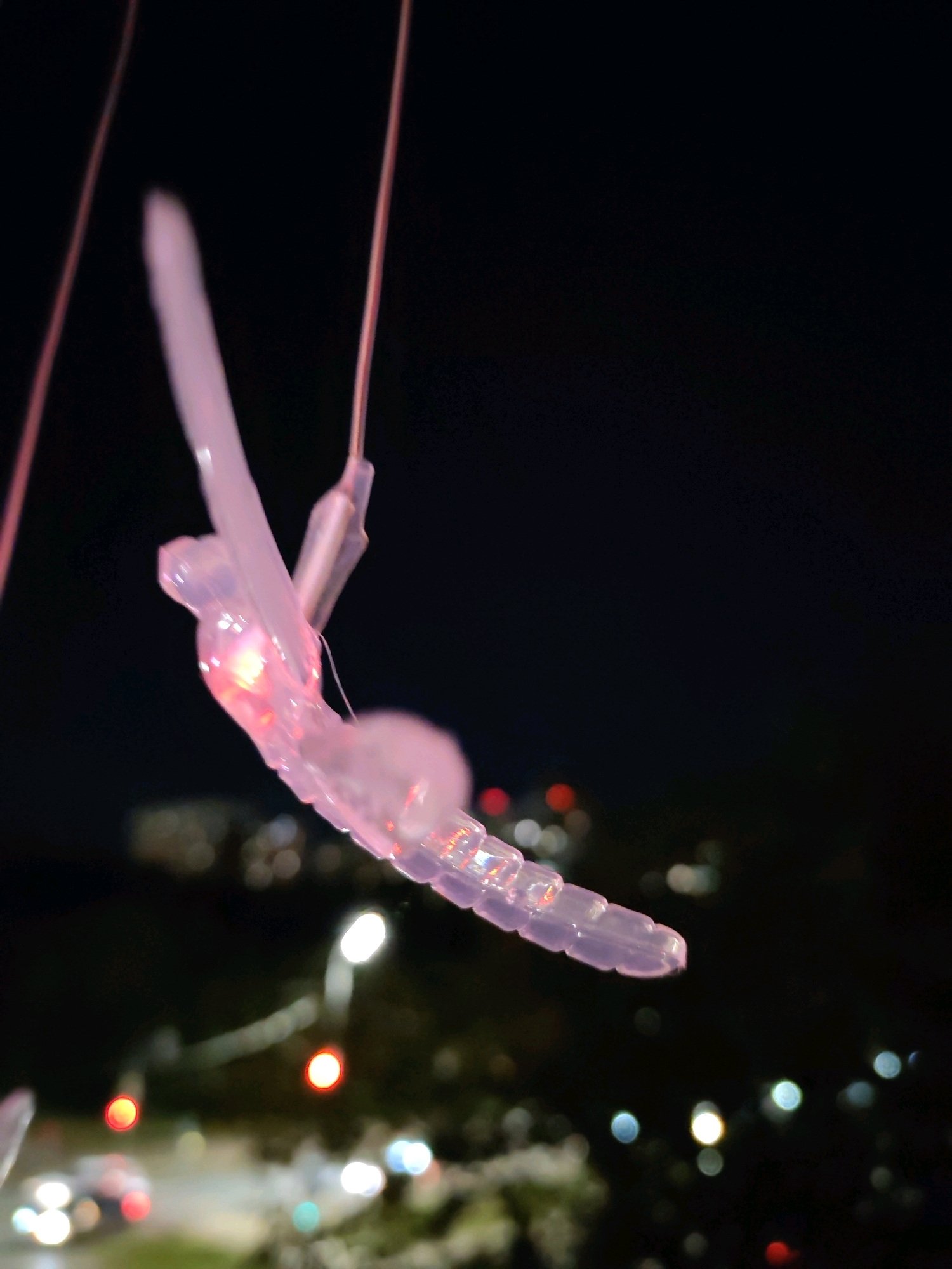 A transparent, pinkish, segmented object, resembling a stylized insect or a piece of light-up garland, is suspended against a dark night sky. The object is illuminated from within, with a distinct pink glow emanating from one section. It appears to be hanging from thin strings or wires. In the background, out of focus, are city lights. These lights are rendered as bokeh circles of various colors, including white, red, and blue, suggesting a busy urban environment. The presence of these lights indicates the photograph was taken at night in a location with artificial illumination. Based on the presence of city lights and the style of buildings visible in the blurred background, the scene is likely an outdoor urban setting in Izhevsk, Russia. No people are visible in the image. The focus is entirely on the illuminated object and the bokeh effect of the background lights.