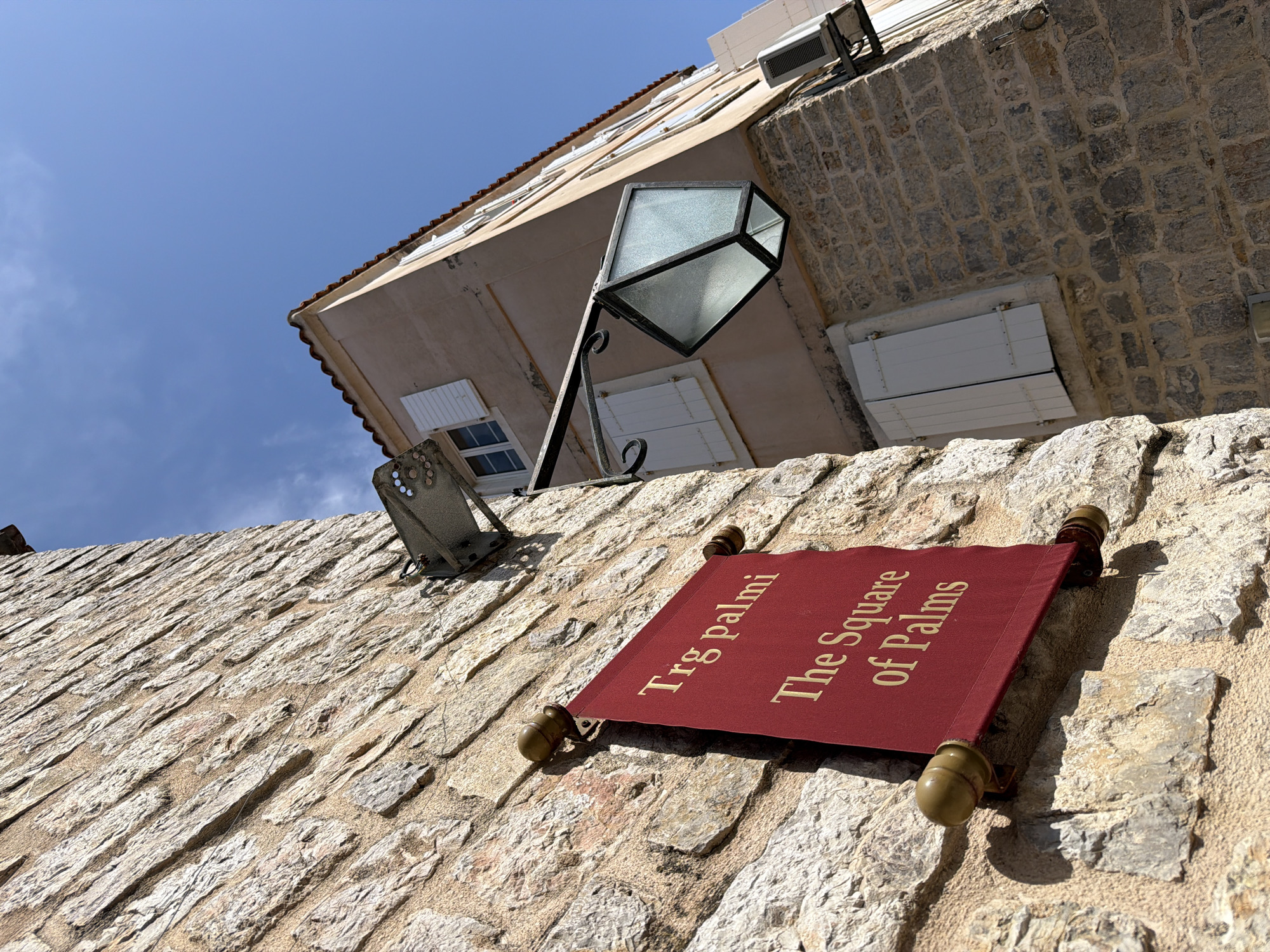The scene is taken from a low angle, looking upwards towards a stone wall in the city of Budva, Montenegro. The wall has a red sign attached to it with the words "Trg palmi The Square of Palms" written in gold letters. A vintage-style street lamp is mounted above the sign. In the background, a multi-story building with light-colored walls, a tiled roof, and white shutters is visible against a clear blue sky with scattered clouds. The buildings and the wall are constructed from rough, weathered stones. An air conditioning unit is affixed to the building's upper section.
