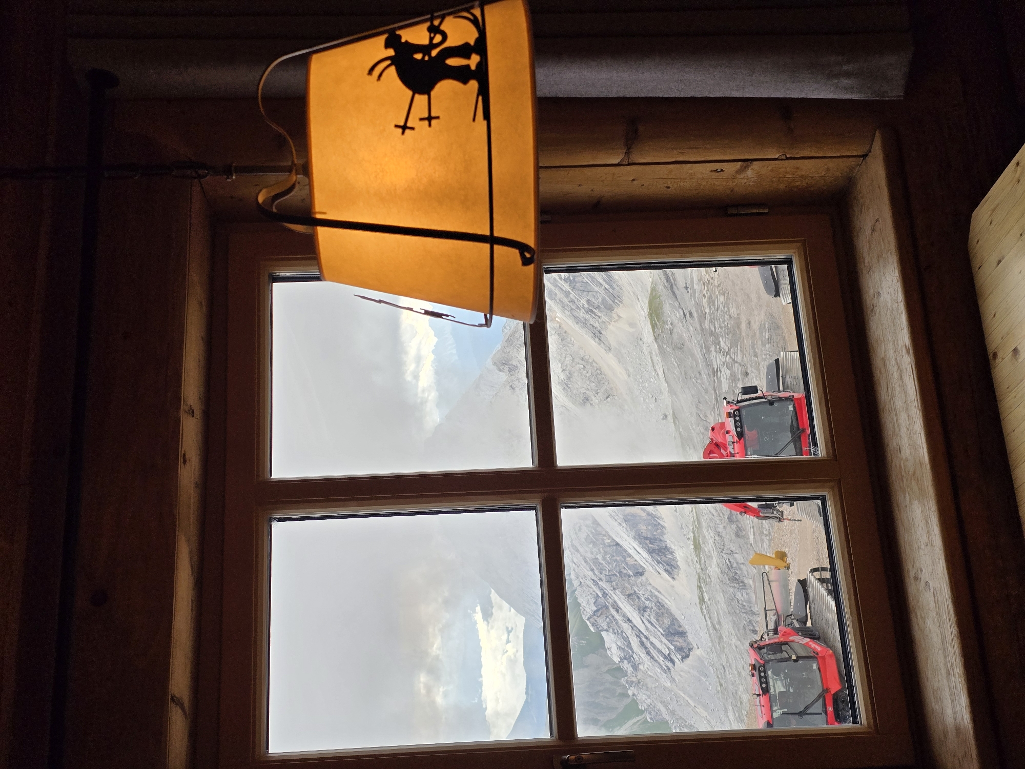The image captures a view through a wooden-framed window, likely from a rustic cabin or guesthouse. The window is divided into four panes, offering a glimpse of a mountainous landscape under an overcast sky. In the foreground, to the right, a lampshade with a silhouette of a skier is visible, lit from within, casting a warm, orange glow. The interior walls are made of dark wood.
Outside the window, the scene is dominated by rugged, rocky mountains, some shrouded in clouds. Below the mountains, two red snow groomers are parked on a gravelly surface. These vehicles are typical of those used for maintaining ski slopes. The presence of snow groomers suggests that this location is in a ski resort area, which, given the context of Garmisch-Partenkirchen, Germany, is highly probable. Garmisch-Partenkirchen is a renowned Bavarian Alps resort town known for its winter sports and dramatic mountain scenery, including the Zugspitze, Germany's highest peak. The weather appears cool and potentially damp, with visible clouds at various altitudes, suggesting conditions conducive to snow or recent snowfall.