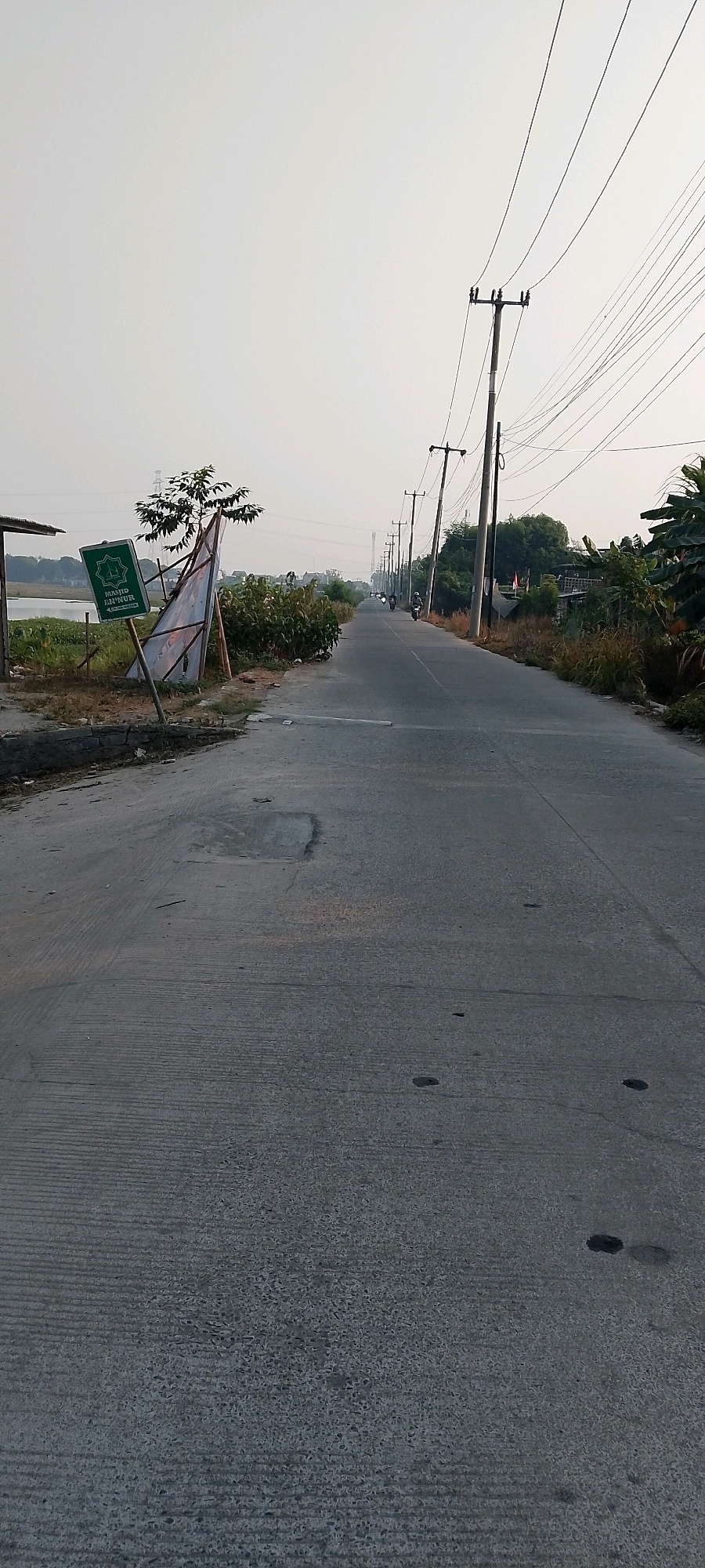 A paved road stretches into the distance, lined with utility poles and electrical wires. On the left side of the road, a green sign with white text and an emblem is propped up by wooden poles. The sign appears to be for a mosque named "MASJID AN-NUR". A body of water is visible behind the sign. Vegetation, including trees and bushes, flanks both sides of the road. Several motorcycles are seen on the road, moving away from the viewer. The sky is overcast and pale. The scene is likely located in a rural or semi-urban area.