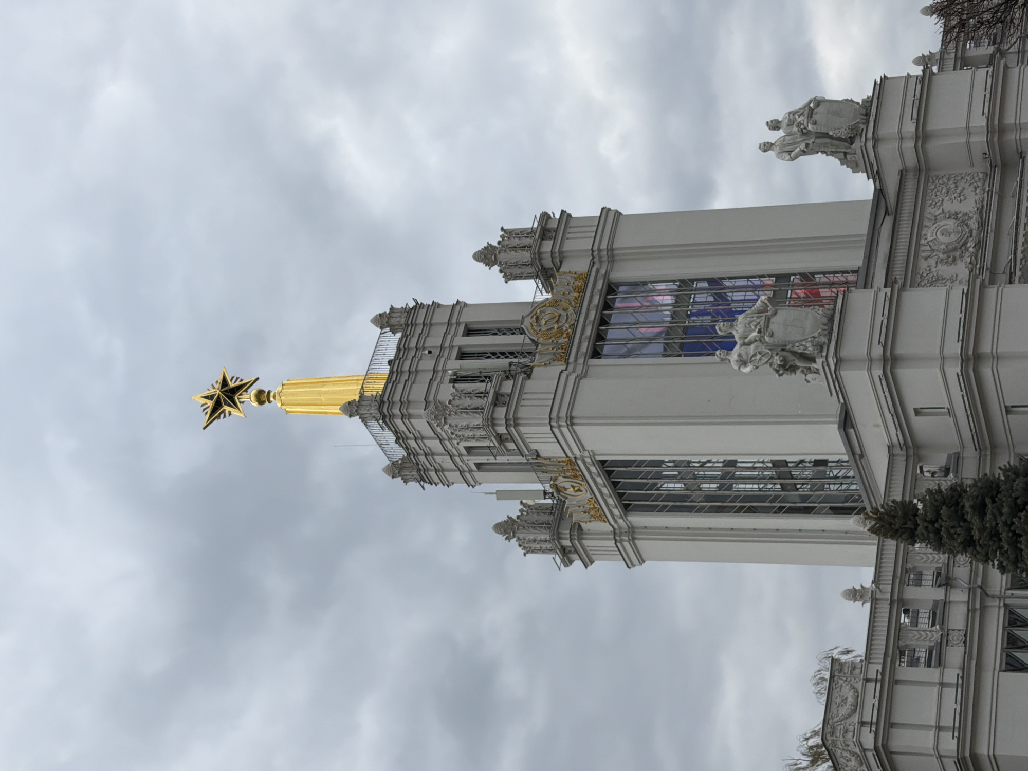 A majestic, ornate building is captured under an overcast, cloudy sky. The image primarily features the upper section of a grand architectural structure, characterized by its light-colored facade (likely white or light grey) adorned with intricate gold detailing. A prominent golden spire culminates in a large, multi-pointed golden star at the building's apex.
The facade displays numerous decorative architectural elements, including ornate moldings, cornices, and balconies. Sculpted figures, appearing as statues, are positioned along the building's exterior, depicting groups of people in heroic poses, some holding shields or other symbolic objects. Large windows, some featuring panes with visible blue and red hues, are integrated into the design.
The sky is filled with diffused grey clouds, indicating an overcast day. A dark green coniferous tree is partially visible at the bottom right, suggesting the presence of landscaping or adjacent structures. The architectural style, with its monumental scale, classical elements, gold accents, and the prominent star, is characteristic of Stalinist-era architecture. Given the location context of Moscow, Russia, this structure is likely a pavilion at the Exhibition of Achievements of National Economy (VDNH).
No human activity is depicted, and no discernible text is visible on the building. The image conveys a sense of grandeur and historical monumentality.
