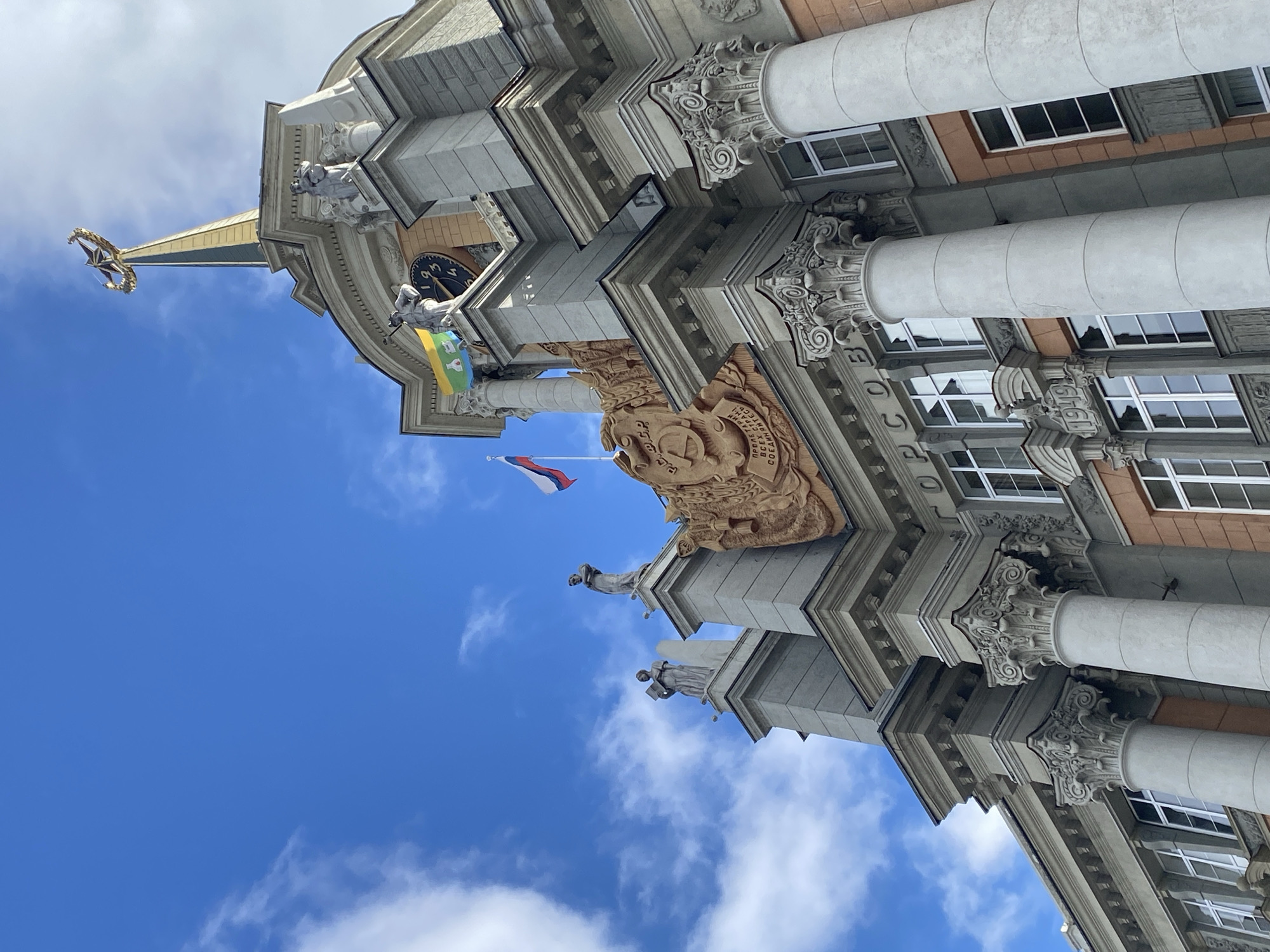 The image captures the City Hall of Yekaterinburg, Russia, from a low-angle perspective, emphasizing its architectural details against a backdrop of blue sky and scattered white clouds. The building showcases a neoclassical style with prominent columns, ornate capitals, and detailed relief carvings. A Soviet-era emblem is visible. A clock tower rises above, topped with a spire and a star. Flags, one Russian and another featuring green and yellow, are attached to the building. Statues adorn the facade at various levels. Windows are regularly spaced along the building's side.