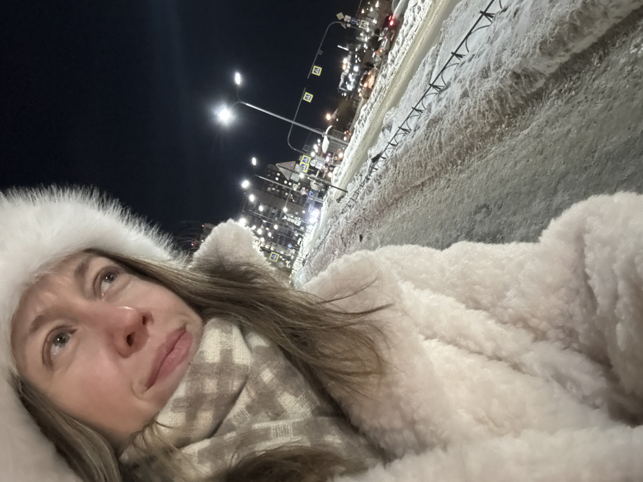 The image depicts an outdoor urban scene at night during winter in Kudrovo, Russia. A female subject is visible on the left, looking towards her right, wearing a white furry hat, an off-white faux fur coat, and a beige and white checkered scarf. The foreground shows a snow and ice-covered pedestrian path. Adjacent to this path is a multi-lane road with active vehicular traffic, indicated by blurred headlights and taillights. The street and sidewalk are illuminated by numerous streetlights on tall poles. A low, dark-colored metal fence runs along the edge of the pedestrian path. In the background, several multi-story buildings are visible, illuminated by both streetlights and internal lights. Multiple yellow pedestrian crossing signs are present on poles along the street.