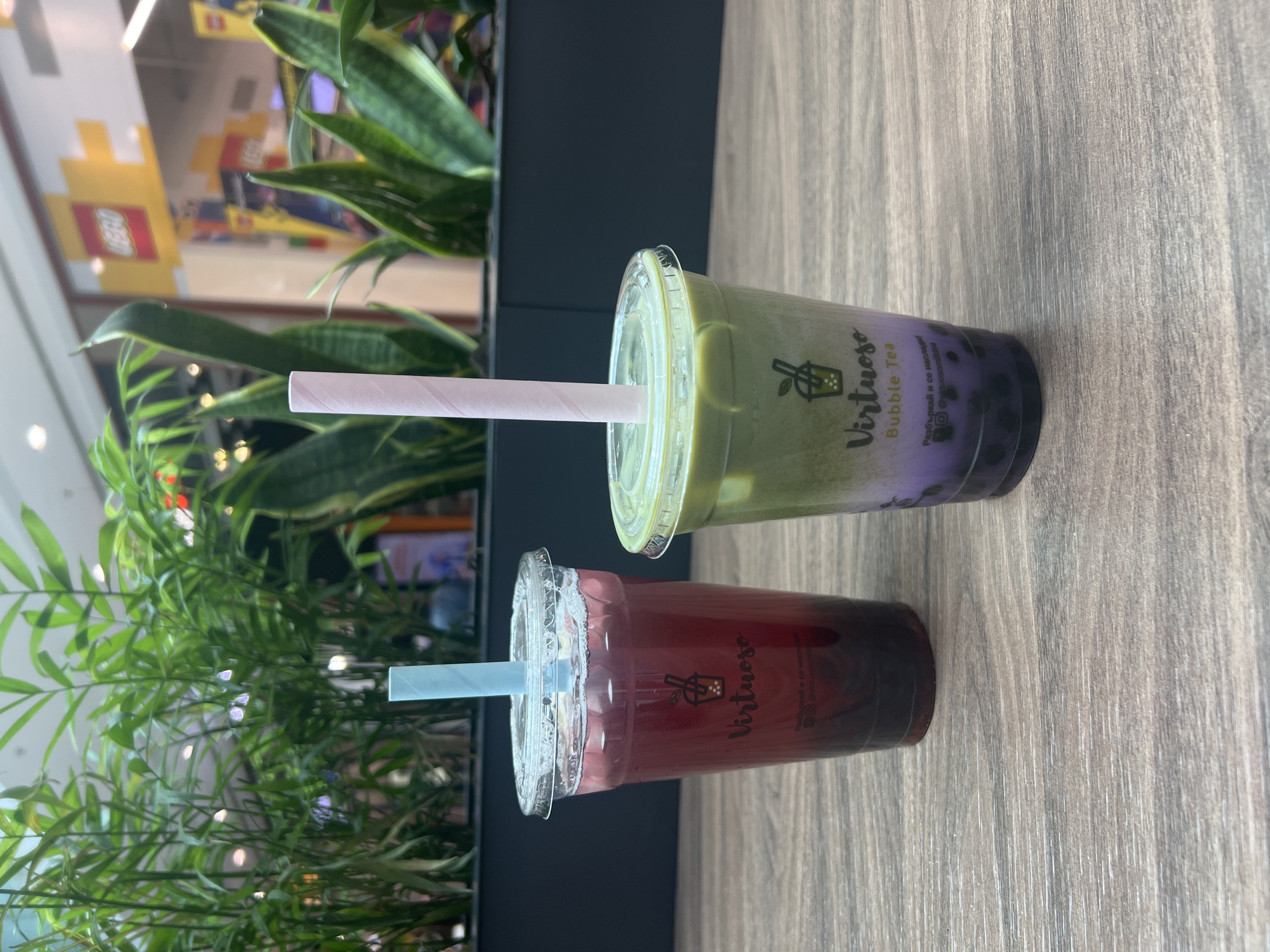 The image shows two plastic cups of bubble tea from "Virtuoso Bubble Tea" sitting on a wooden table or counter. One cup contains a green tea mixture with purple tapioca pearls at the bottom and a pink straw. The other cup contains a red tea mixture and a light blue straw. Green plants surround the drinks on one side. The background features a building interior with a colorful "LEGO" sign visible. This scene is located in Sofia, Bulgaria.