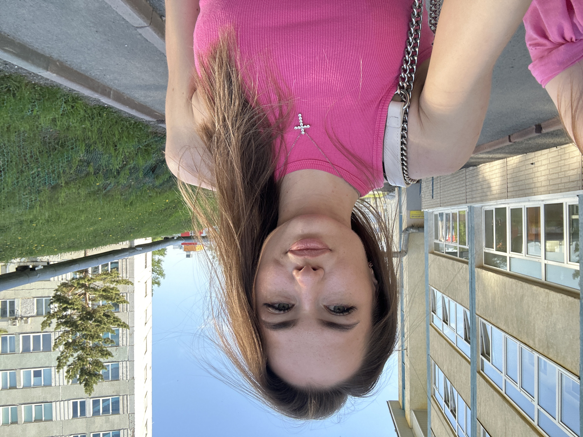 The media file is an outdoor portrait, rotated 90 degrees clockwise, featuring a young woman. She is positioned centrally, appearing to look directly into the camera with a neutral expression. She has long, straight dark brown hair, fair skin, and is wearing light makeup, including defined eyelashes and glossy lips. She wears a fitted, ribbed, sleeveless pink top. A silver necklace with a prominent studded cross pendant rests on her chest. Part of a white handbag with a silver chain strap is visible over her right shoulder.
The background shows an urban or suburban environment under a clear blue sky, suggesting sunny weather and daytime. To her left (viewer's right in correct orientation), a large, multi-story building with numerous windows is visible, partially fronted by a tall, green-leafed tree. To her right, another building with windows and a section of brick facade can be seen. The foreground includes patches of green grass, a gray curb, and paved ground, indicative of a street or park area adjacent to buildings. No specific location cues or text are visible, and the overall mood conveyed is calm.