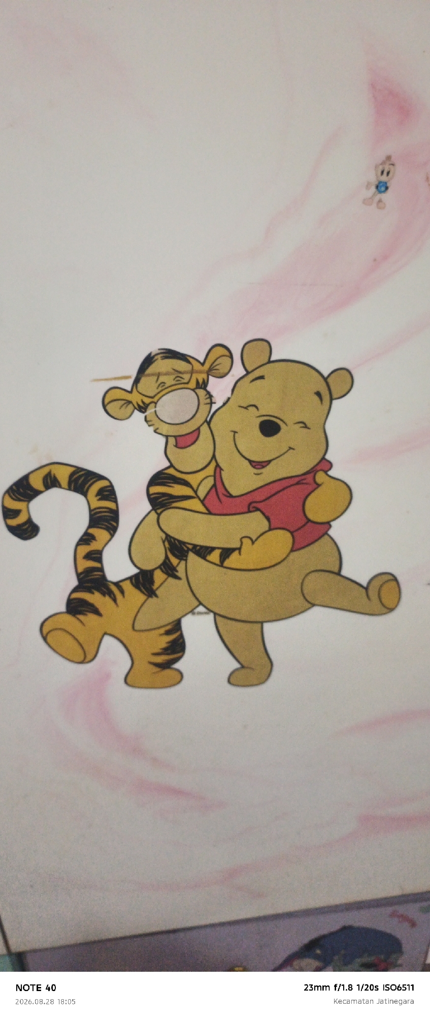 The image features a cartoon illustration of Winnie the Pooh and Tigger embracing. Winnie the Pooh, a yellow bear, is wearing a red shirt and is positioned on the right, hugging Tigger, a striped orange tiger. Tigger is on the left, with his arms around Winnie the Pooh. The background is a soft pink and white marble pattern. At the bottom of the image, overlaid text indicates "NOTE 40 2026.08.28 18:05" on the left and "23mm f/1.8 1/20s ISO6511 Kecamatan Jatinegara" on the right. The text suggests the photograph was taken in Jatinegara, East Jakarta, Indonesia.