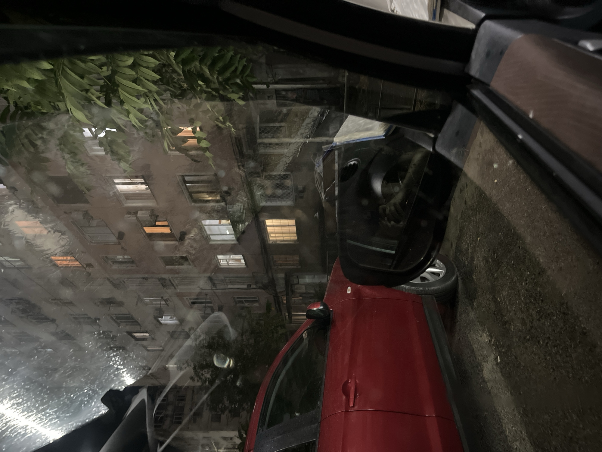 The image captures a nighttime scene through the window of a vehicle, revealing a residential building with lit windows in Poti, Georgia. A red car is parked to the left, its side visible. The foreground is dominated by the interior of the car, including the side mirror which reflects the scene. Raindrops or water droplets are visible on the exterior surface of the vehicle's window, creating a blurred and streaky effect. The building appears to be multi-story with multiple windows, some of which are illuminated with warm light. Lush green foliage is present on the right side of the frame, partially obscuring the building. The overall atmosphere is moody and captures a glimpse of urban life at night in Poti.