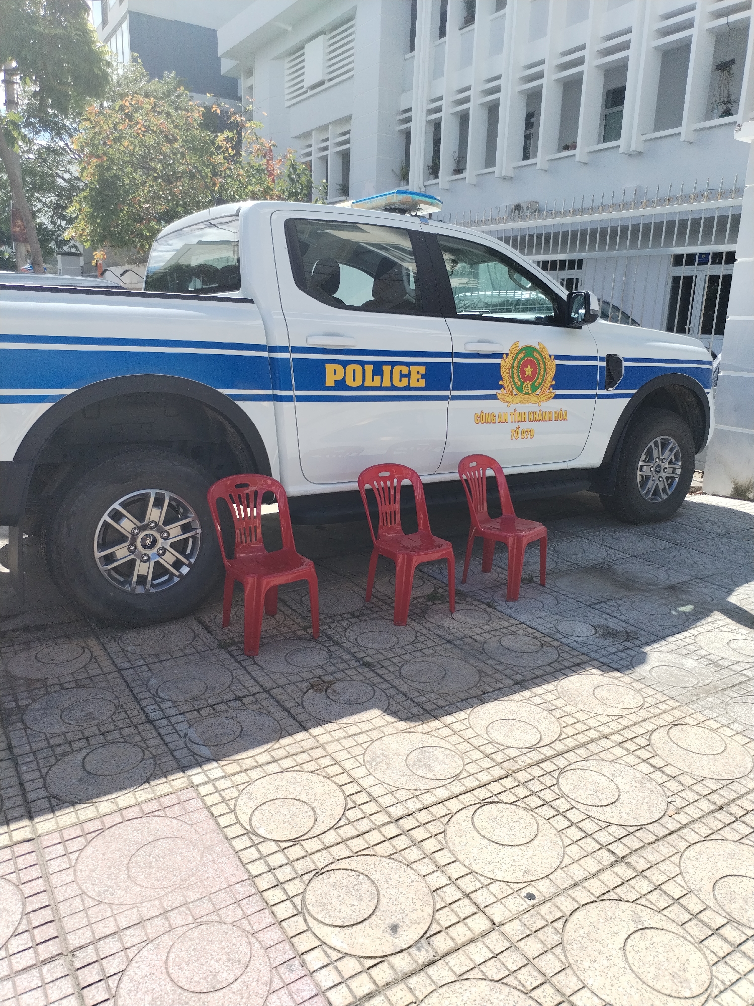 A bright outdoor scene captures a white police pickup truck parked on a patterned paved surface. The truck, oriented towards the right, has a prominent blue stripe along its side, with "POLICE" written in large yellow capital letters. Next to "POLICE" is a gold-colored emblem featuring a star and a wreath. Below the emblem, Vietnamese text reads "CÔNG AN TỈNH KHÁNH HÒA" (Public Security of Khanh Hoa Province) and "TỔ 079" (Group 079).
In front of the truck's side, three identical bright red plastic chairs are neatly lined up. The ground is paved with square tiles, each containing a circular pattern, and is partially shaded, suggesting a sunny day with strong sunlight.
In the background, a light-colored, multi-story building with numerous windows and a white facade is visible, possibly an official building given the police vehicle's presence. Lush green trees and foliage are seen behind the building and to the left of the frame, providing a natural backdrop. The overall setting appears to be an outdoor area, likely in Nha Trang Ward, Vietnam, near a public security office. No people are visible, and there is no apparent activity.