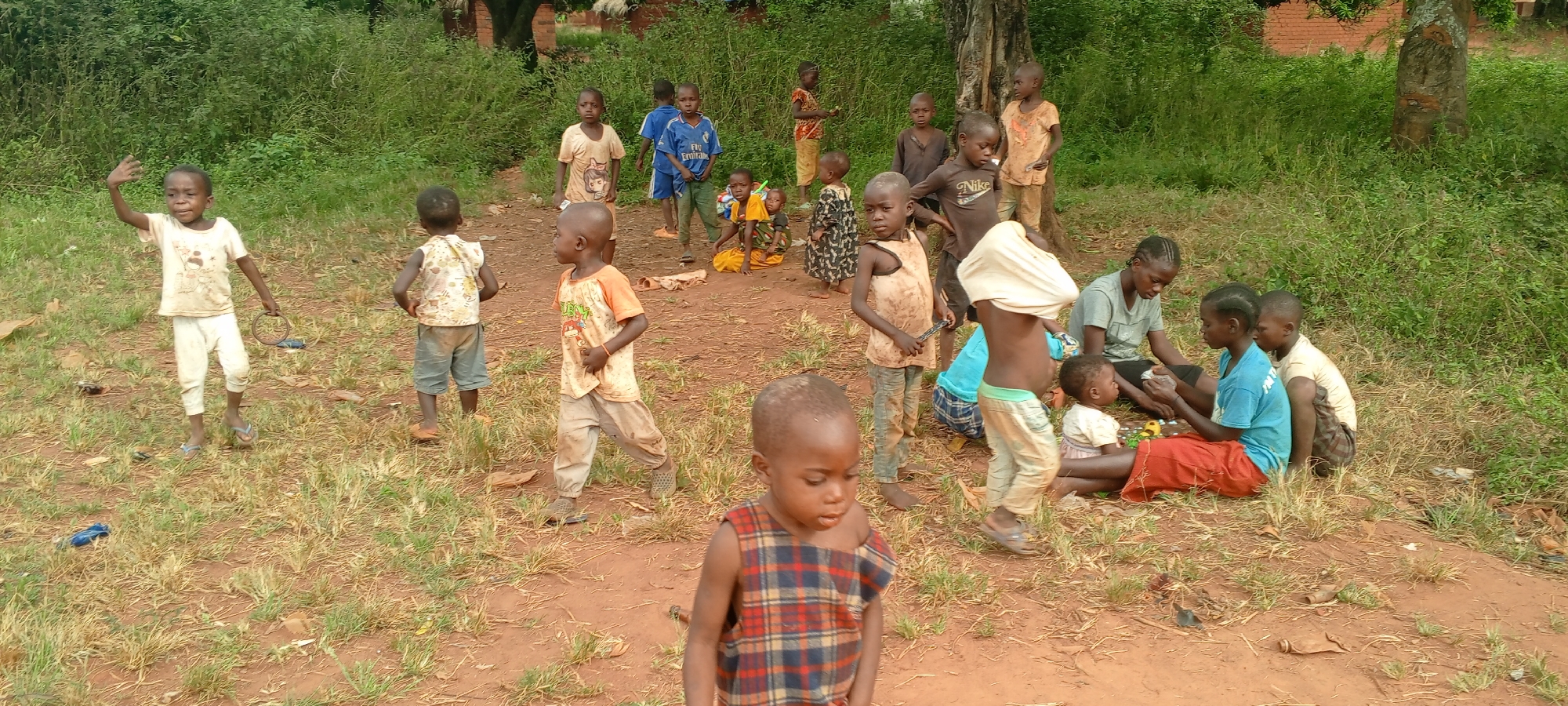 In Ngalula, Democratic Republic of the Congo, a group of children are in an outdoor area with sparse grass and red soil. Some children stand and walk in the open space while others gather near adults sitting on the ground. The children wear various clothing, including t-shirts, pants, dresses, and sandals. Adults wear t-shirts and skirts, one holds a baby, and they appear to be interacting with small objects on the ground. A tree and dense vegetation are in the background.