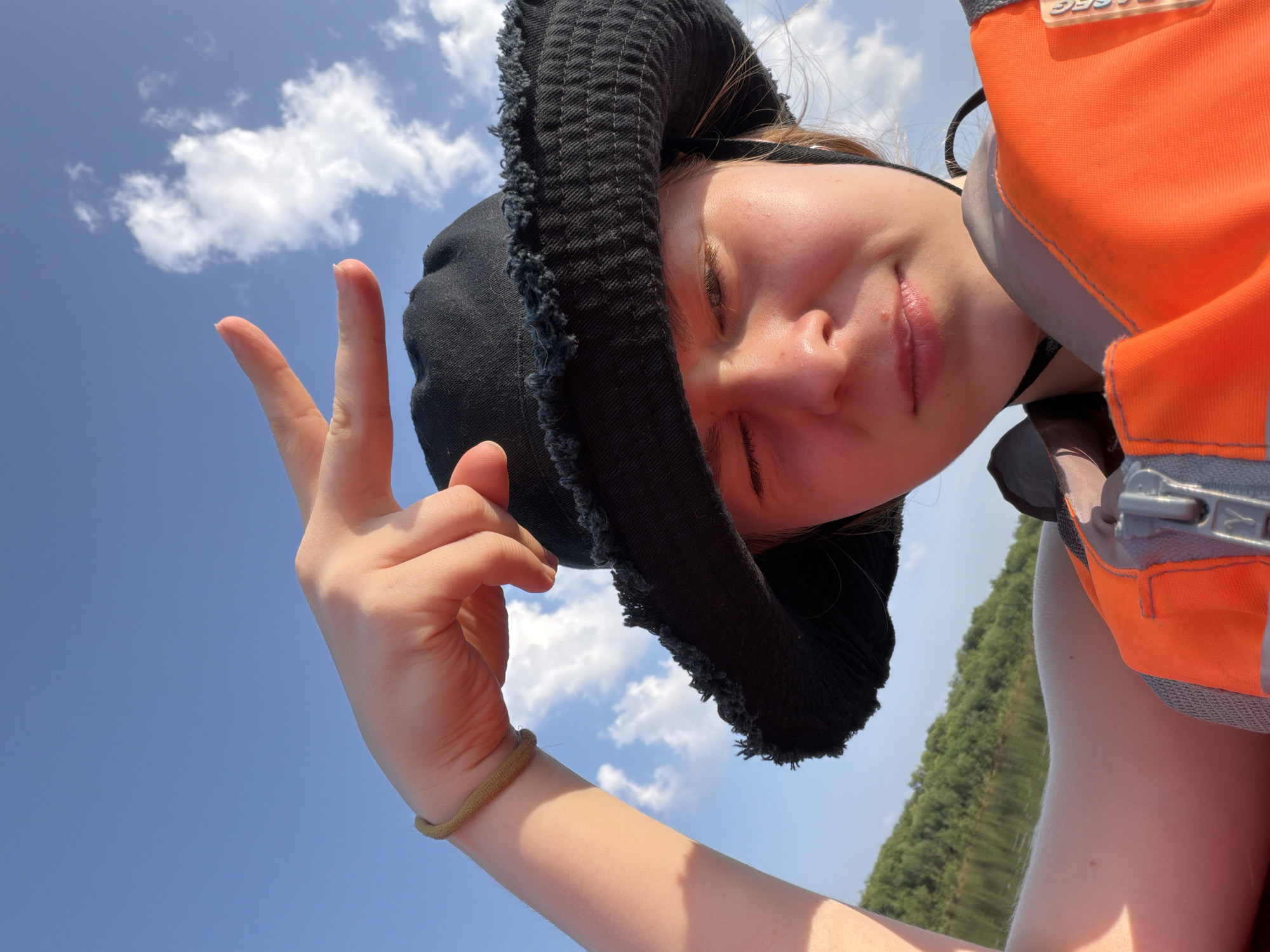 A young person wearing a black bucket hat and an orange life vest is taking a selfie. They are holding up their hand in a peace sign gesture. The background shows a blue sky with white clouds and a green, tree-lined body of water, suggesting an outdoor activity likely on a lake or river in Kazaevo, Russia. The sun is casting a warm glow on the person's face.