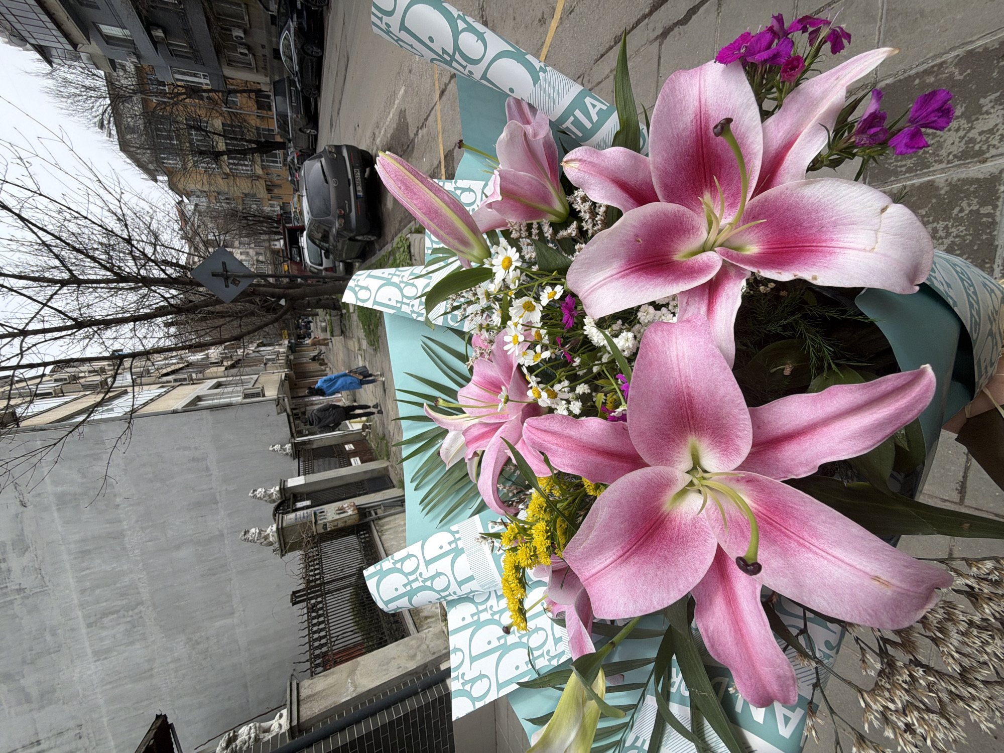 The image shows a bouquet of flowers in the foreground, with pink lilies being the most prominent. There are also smaller white and yellow flowers, and some purple flowers. The bouquet is wrapped in light blue paper with a repeating pattern of "Dior" logos.
The background depicts an urban street scene. There are buildings on either side of the street. The buildings are of various heights and architectural styles, typical of older European cities. A bare tree is visible on the left. There are cars parked along the street.
Based on the architecture and street layout, the location could potentially be in Varna, Bulgaria.