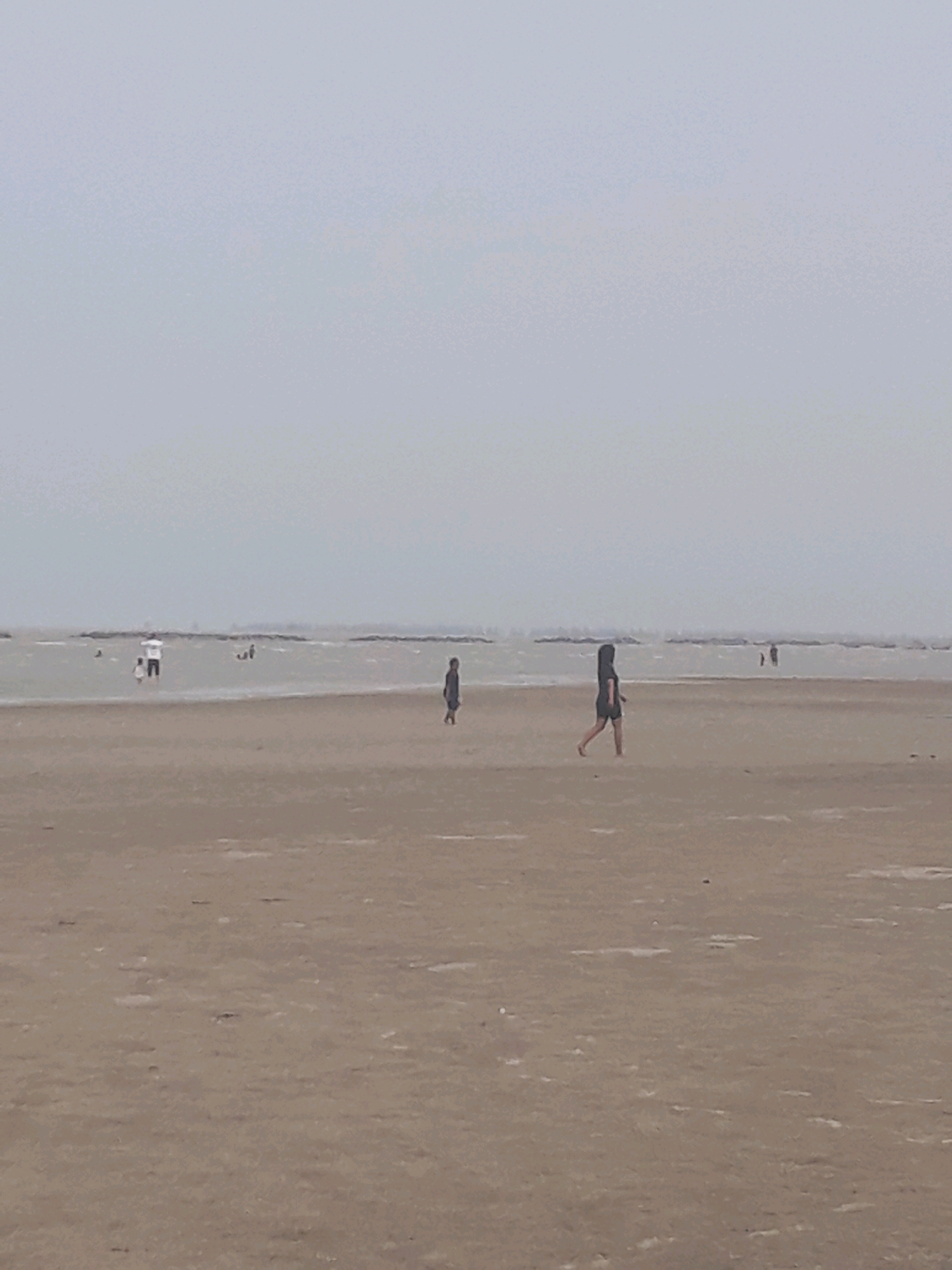 The image shows a wide, flat, sandy beach under an overcast sky. Several people are visible in the distance, wading in the shallow water or walking on the wet sand near the shoreline. In the foreground, a lone individual, appearing to be a woman with dark hair, walks across the dry sand from left to right. The water is calm, with gentle waves breaking near the shore. A distant, hazy horizon line suggests the presence of land or structures far across the water. The overall scene is muted and subdued due to the cloudy weather.