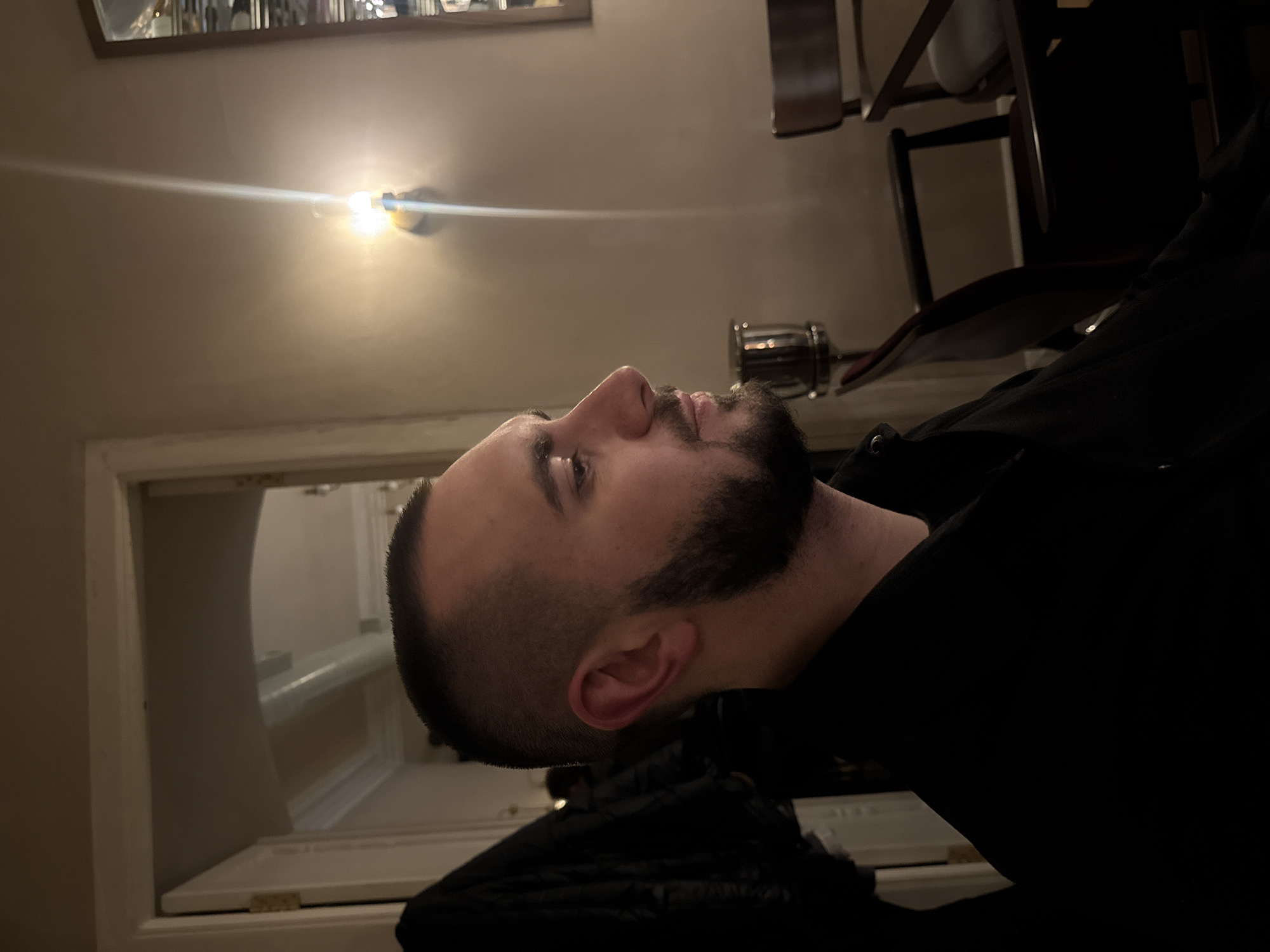 A low-angle, interior shot shows a man in Yerevan, Armenia. The man has short, dark hair, a beard, and is wearing a black jacket. He is looking upwards. A light fixture is visible on the ceiling. In the background are parts of chairs, a mirror, and a doorway.