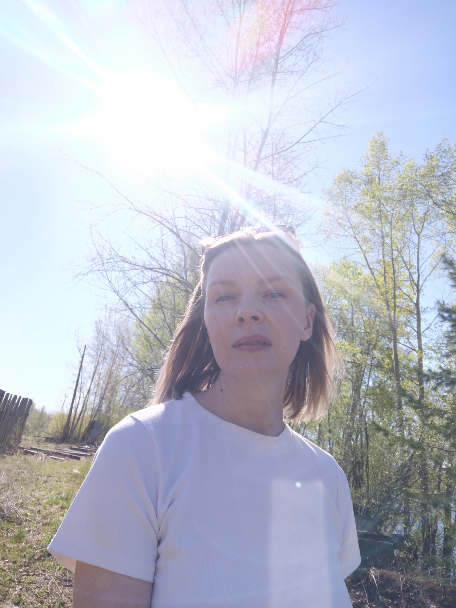 A medium shot shows a fair-skinned, light-haired woman wearing a white t-shirt, posing outdoors. The background consists of trees, some with bare branches and others with light green foliage. A section of a weathered wooden fence is visible on the left. The sky is blue with a bright sun creating significant lens flare. Based on geographic location in the city of Chaikovsky, Russia, the vegetation suggests a park or natural area near the Kama River.