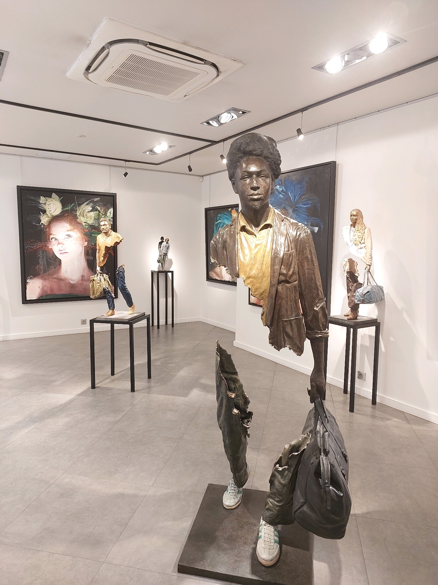 This image shows the interior of an art gallery in Saint-Paul-de-Vence, France. The gallery features several sculptures and paintings. In the foreground, a life-sized bronze sculpture of a man is displayed. The sculpture is partially fragmented, with the man's torso and legs visible, holding a black duffel bag. He is wearing a yellow shirt and a brown leather jacket, with white sneakers. To the left, a painting of a woman's face with flowers is on display. To the right, another bronze sculpture of a woman is displayed on a pedestal, also partially fragmented, holding a blue bag. The gallery has white walls and a grey tiled floor, with track lighting on the ceiling.
