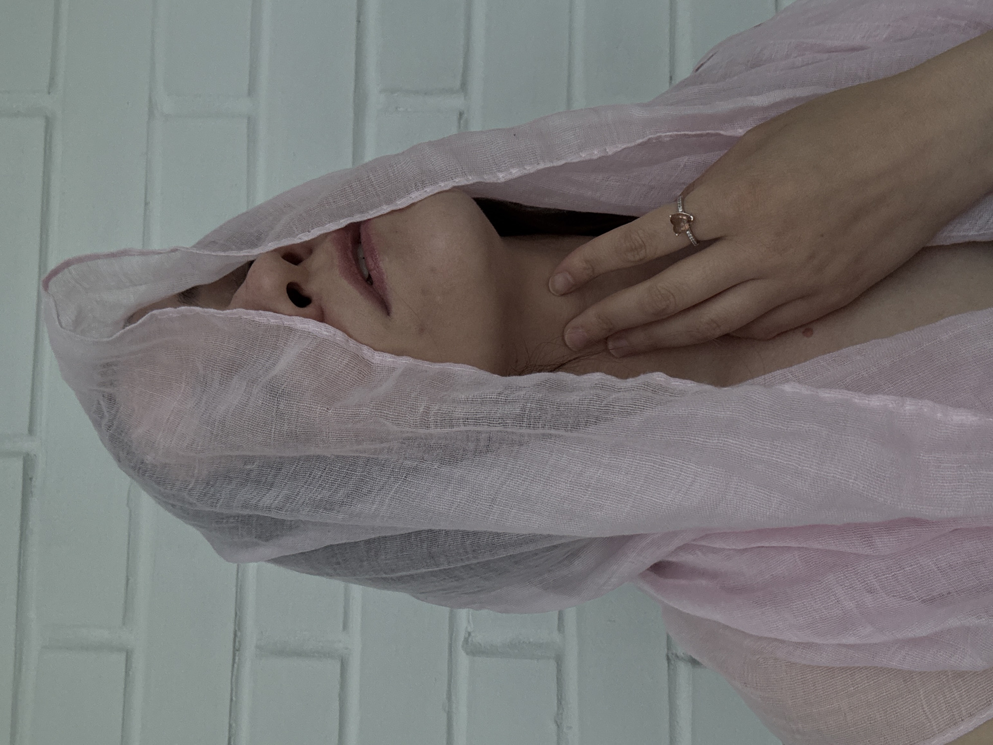 This portrait-oriented photograph, originally captured at a 90-degree clockwise rotation, depicts a young adult individual, partially enveloped by a light pink, sheer, loosely woven fabric, likely a scarf or veil. The subject's chin, mouth (slightly open, revealing a hint of teeth), and nose are visible, while their eyes and forehead are obscured by the draping fabric. A hand, adorned with a ring featuring a light-colored central stone (possibly heart-shaped) and smaller accent stones, gently rests on the subject's neck. The fabric creates soft folds around the visible parts of the face and upper body. The background is a light-colored brick wall, likely painted white or a very pale grey, showing the distinct pattern and texture of bricks. The lighting is soft and even, suggesting an indoor environment or diffused light. The pose and partially obscured face convey a sense of contemplation or serenity. There is no visible text in the image. The location context for the image is Chaikovsky, Russia.