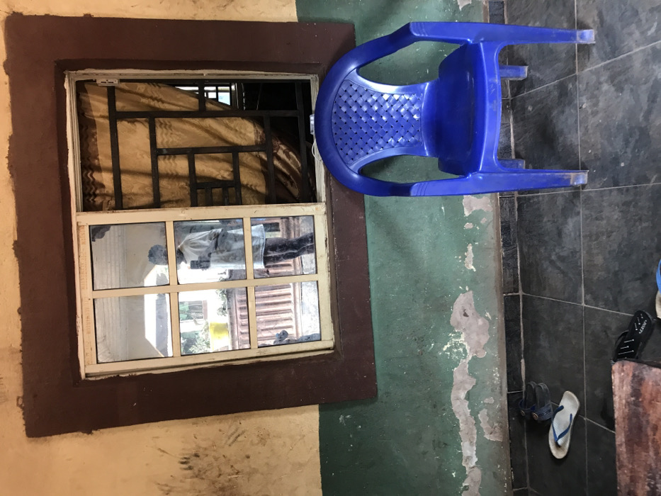 A window with brown trim has metal bars and a tan curtain partially obscuring the view. The window reflects objects from the outside. Next to the window sits a blue plastic chair. The wall is painted two colors: green at the bottom with peeling paint, and yellow at the top. Two pairs of sandals and a pair of flip-flops are on the tiled floor.