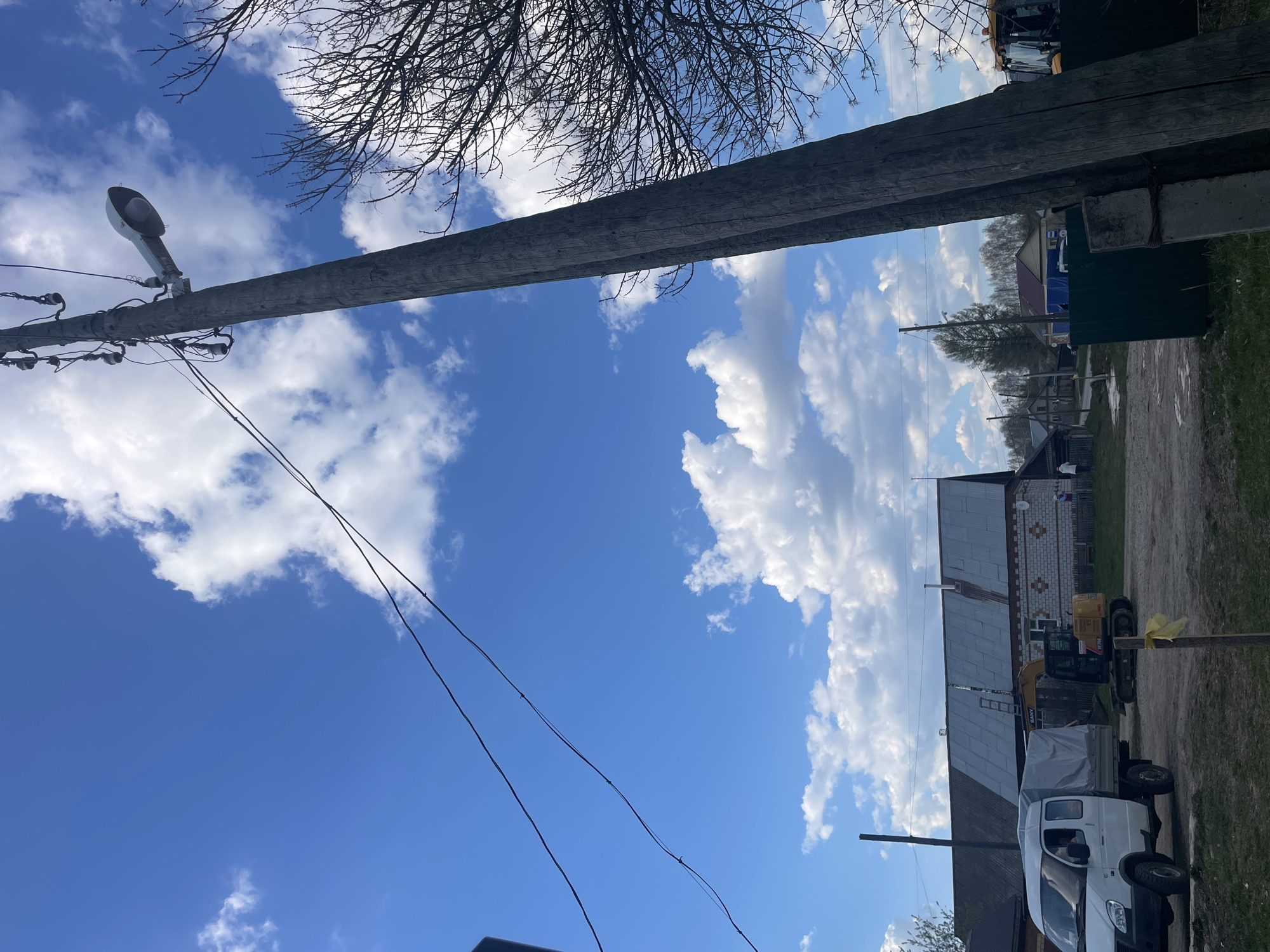 The image shows a rural scene in Поедуги, Russia, with a focus on a weathered wooden utility pole carrying a street lamp and overhead wires. Above the pole, bare tree branches are visible against a backdrop of blue sky with scattered white clouds. In the background, traditional Russian houses are lined along a dirt road, with a white van and a yellow excavator parked nearby. The sky is predominantly blue with puffy clouds. Mud is visible on the road.