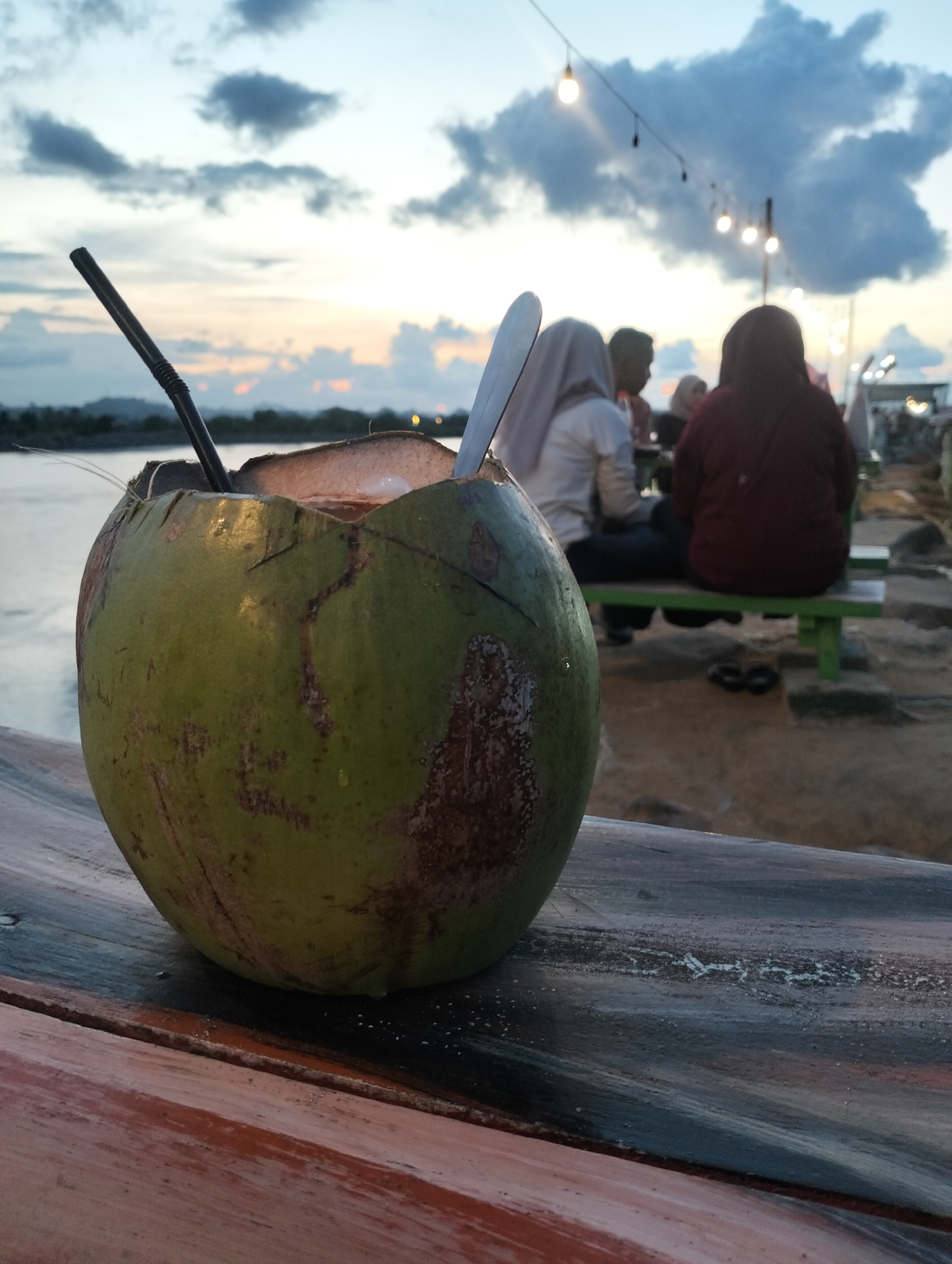 On a wooden surface, a young coconut is situated with a straw and spoon inserted. In the background, a group of people sit on a bench. Above them are strung lights. The scene is set along a body of water, presumably at a coastal location in Lhokseumawe, Indonesia, given the lighting and general atmosphere.