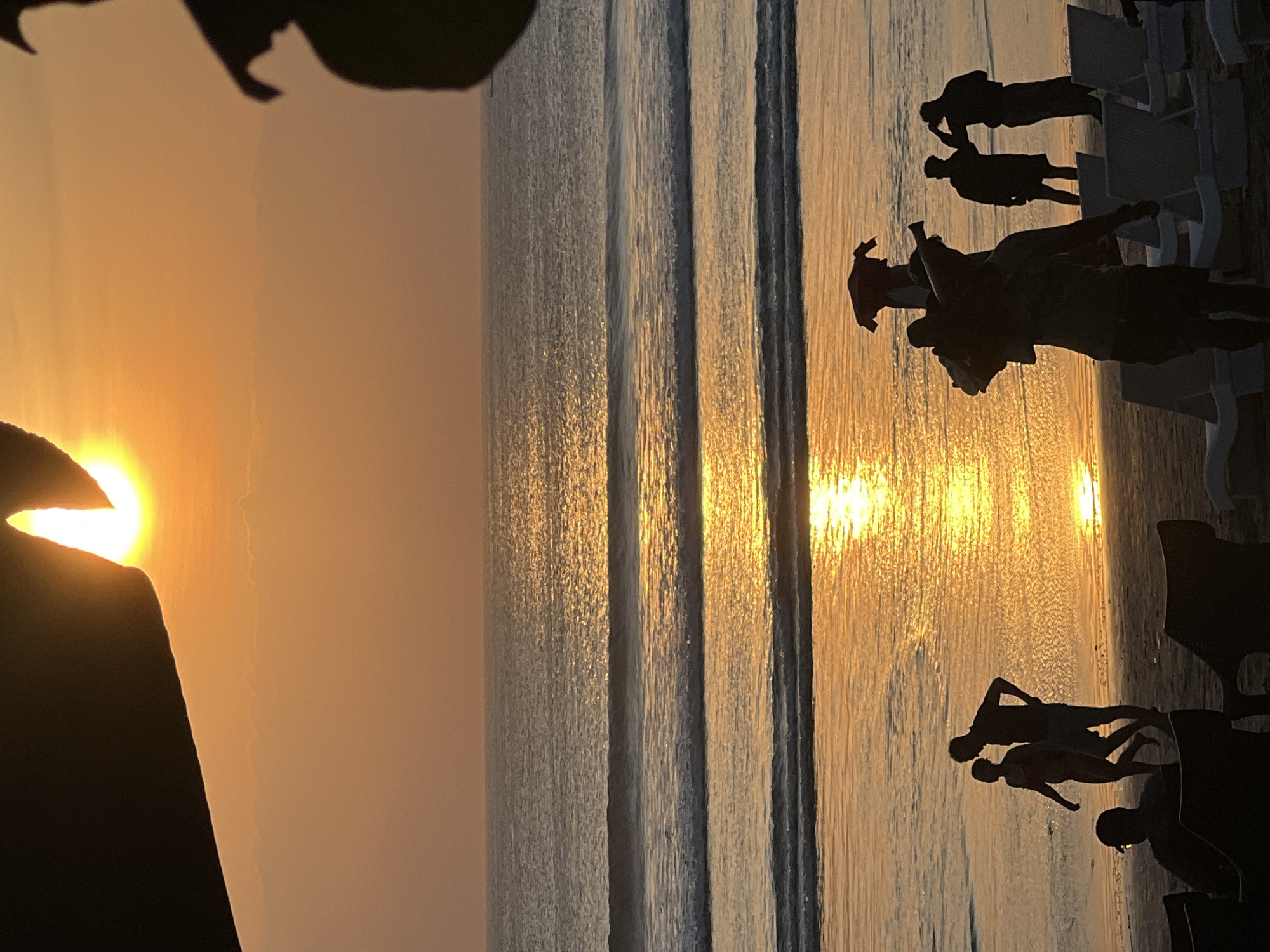 This is a photograph taken at sunset on Kuta Beach in Kuta, Indonesia. The sun, partially obscured by what appears to be a dark silhouette of an object in the foreground, is low on the horizon, casting a warm, golden glow across the sky and the ocean. Gentle waves roll onto the sandy shore, their surfaces reflecting the vibrant light.
Several silhouetted figures are visible on the beach. In the mid-ground, a couple walks hand-in-hand along the water's edge. Further along the beach, a group of people are gathered, some appearing to be engaged in conversation. In the foreground, lounge chairs are set up on the sand, some occupied by silhouetted individuals. The overall scene is tranquil and evokes the serene atmosphere of a beach at dusk.