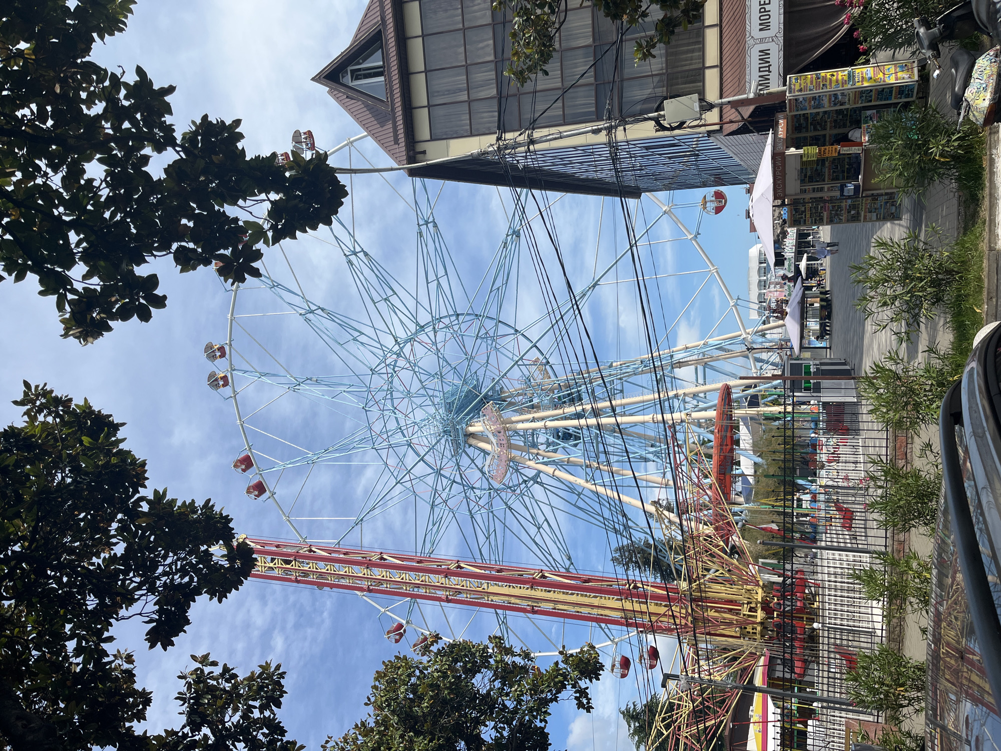 This image shows an amusement park in Sochi, Russia, on a sunny day with a partly cloudy sky. A large Ferris wheel with blue spokes and red gondolas dominates the center of the frame. To the left of the Ferris wheel is a tall, red and yellow amusement ride that appears to be a drop tower. In the foreground, there is a black fence with metal posts, behind which are various amusement park attractions including what looks like a carousel with red horses. Several people are visible walking around the park. To the right of the Ferris wheel is a multi-story building with a brown, sloped roof and large windows. The building has signs in Russian, one of which reads "МИДИИ МОРЕ" (Midi Mari). There are also several kiosks and umbrellas set up in the park, likely for selling goods or services. Trees with dark green leaves frame the top of the image. A parked car is partially visible in the lower foreground, and a scooter is parked near the building on the right.