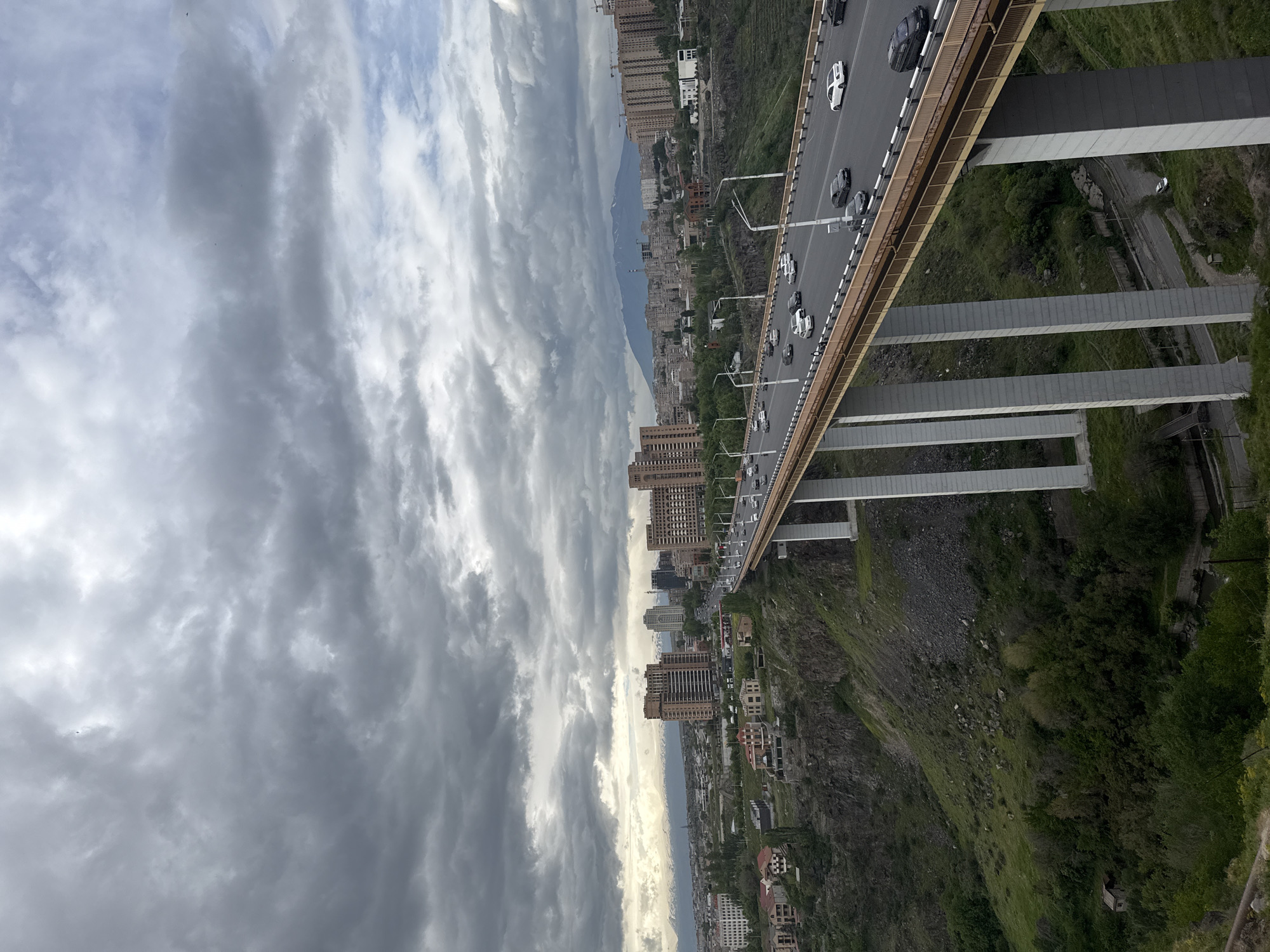 The image captures a high-angle, landscape-oriented view of the city of Yerevan, Armenia, featuring the Davtashen Bridge. The bridge, with multiple vehicles traveling across it, is supported by vertical and horizontal concrete pillars. Dense cloud cover dominates the left side of the frame, contrasting with the cityscape on the right. Below the bridge, a steep, rocky hillside is covered with green vegetation. Tall residential buildings are visible along the city's edge. A road runs parallel to the bridge at a lower level, nestled into the hillside.