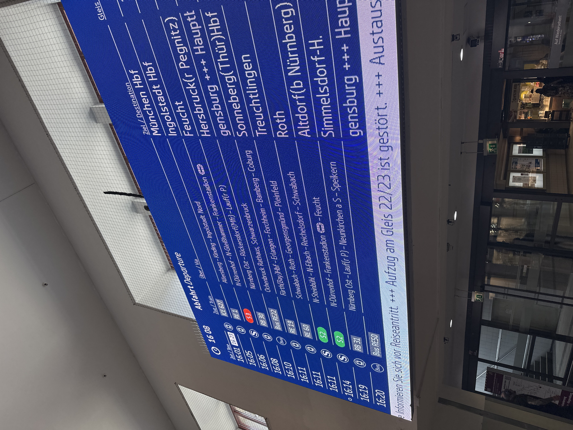 The image displays an indoor scene at a train station, likely in Nuremberg, Germany, as indicated by place names like "Nürnberg Ost" on the departure board. The focal point is a large digital departure board with a blue background and white text, listing train and bus information.
The board shows departure times ranging from approximately 16:01 to 16:20 in the afternoon. Destinations include "München Hbf," "Ingolstadt Hbf," "Feucht," "Hersbruck(r Pegnitz)," "Sonneberg(Thür)Hbf," "Treuchtlingen," "Roth," "Altdorf(b Nürnberg)," and "Simmelsdorf-H." Various train and bus services are listed, such as "ICE 627," "RE 1," "S 3," "RB 30," "Bus RE22," "RE 19," "RE 60," "S 2," "RB 31," and "Bus RE50." Intermediate stops ("Über/Via") are also detailed, for example, "Allersberg-Kinding - Ingolstadt Nord" and "N-Dürrenhof-N-Gleißhammer - Frankenstadion." A crucial message at the bottom of the board informs passengers: "Informieren Sie sich vor Reiseantritt. +++ Aufzug am Gleis 22/23 ist gestört. +++ Austaus" (Please inform yourself before travel. +++ Elevator at platform 22/23 is out of order. +++ Replacement...).
To the right of the departure board, there's a commercial area. A shop, identified as "Brezen Kolb" by its sign, has a glass entrance. Above the shop entrance, an illuminated green "EXIT" sign is visible. A person, with their back partially turned, stands near the shop. The ceiling features a grid-like pattern with integrated lighting fixtures. The scene depicts a typical busy atmosphere of a railway station concourse.