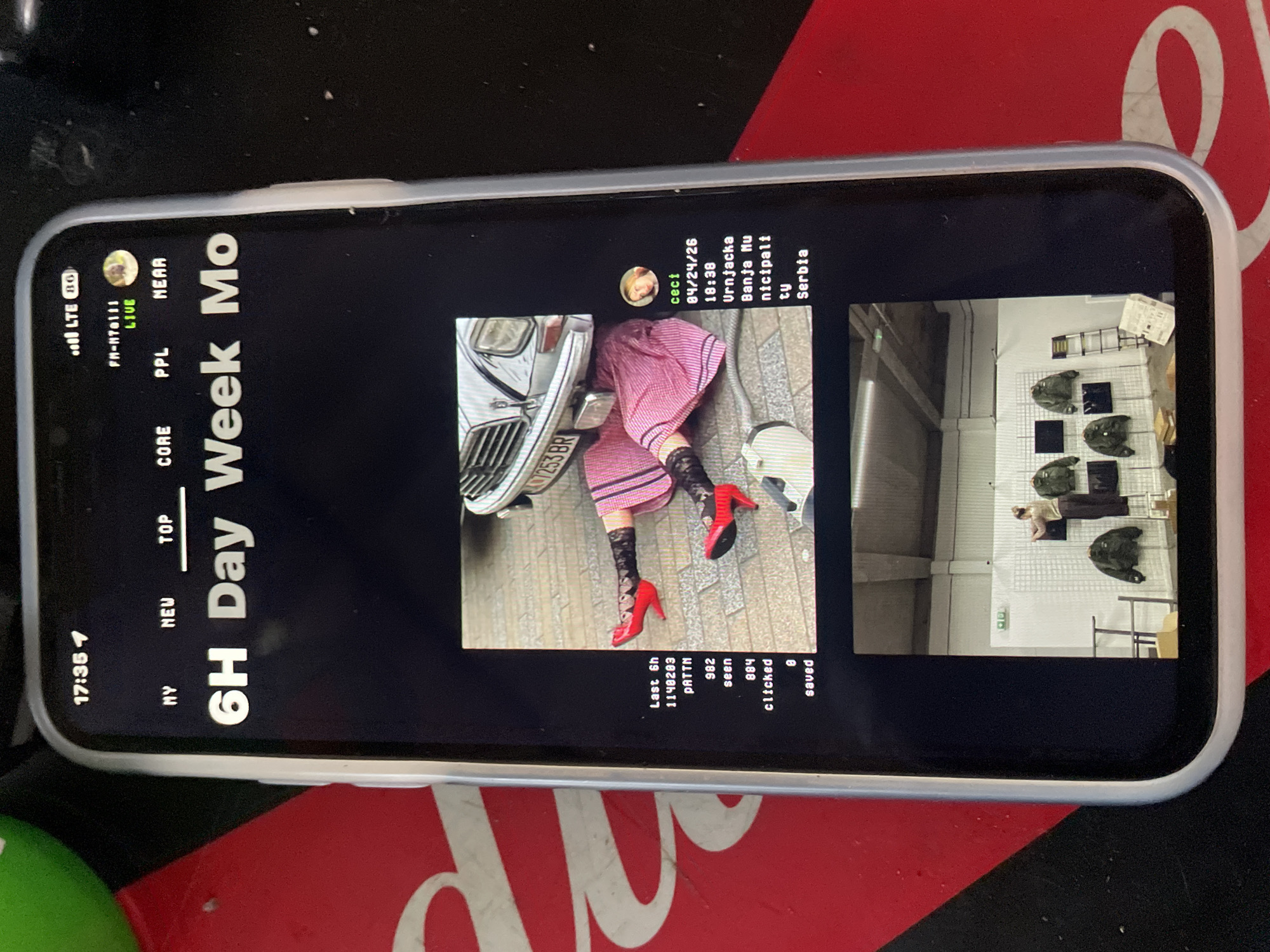 The image shows a mobile phone displaying an interface with two images and associated metadata. The first image shows a woman in a pink dress and red high heels standing near a car with a license plate that appears to include the characters "07253 BR." The second image shows what appears to be a clothing store interior with garments displayed on a rack. Text overlays include "Last 6h 1140203 PATTN 982 seen 884 clicked 8 saved" and "ceci 04/24/26 18:38 Urnjacka Banja Mu nicipali ty Serbia." The phone interface also displays "6H Day Week Mo" along with other status indicators like "LTE," "FM-N70111," "LIVE," "NY," "NEW," "TOP," "CORE," "PPL," and "NEAR." Time displayed is "17:35 1." The phone is lying on a surface that appears to be red and black. There is no indication that the scene is in Jos, Nigeria.