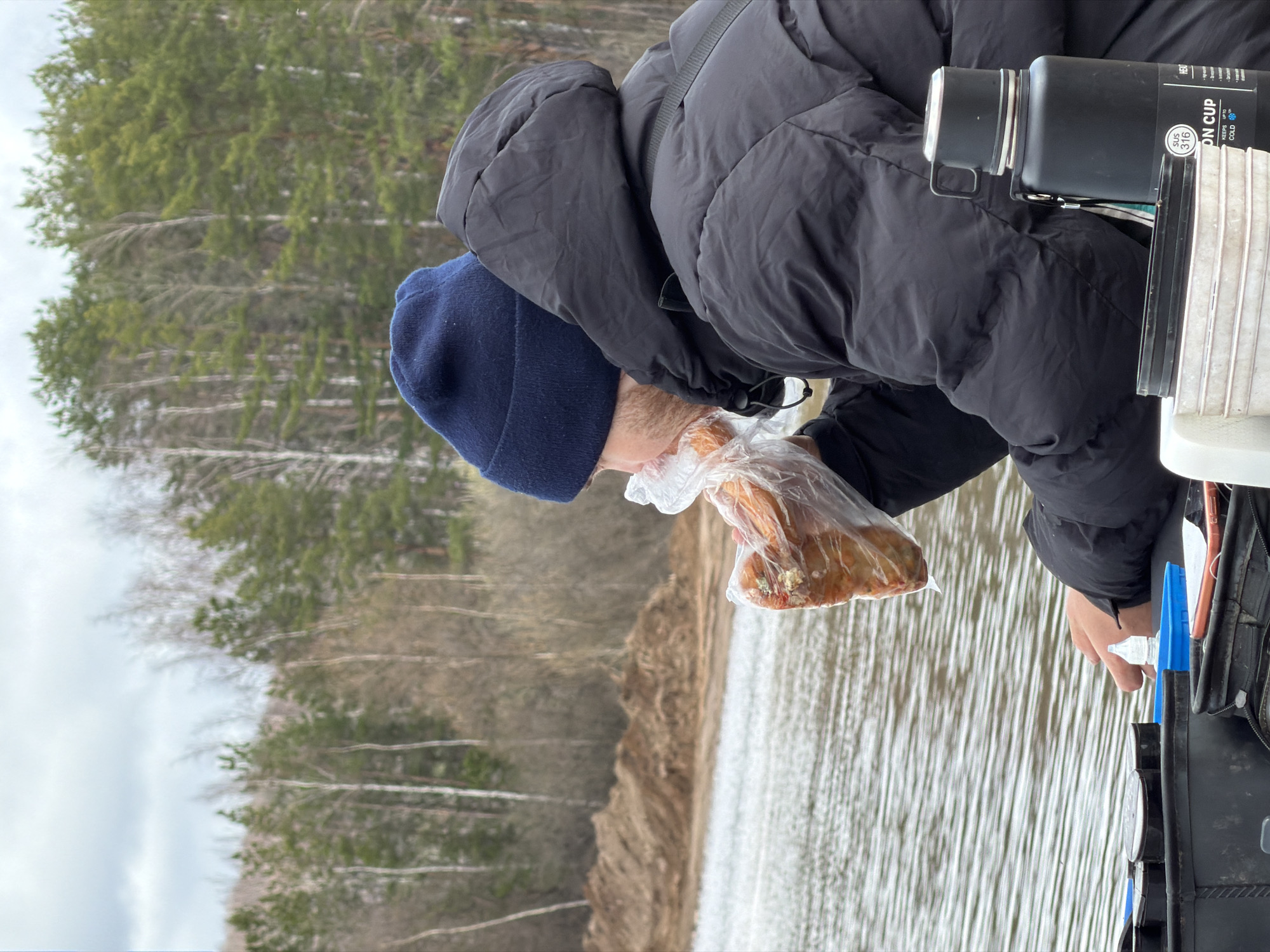 A man wearing a black puffer jacket and a blue beanie is holding a clear plastic bag containing pieces of bread. The man is standing near a body of water, likely a lake or river, as trees are visible in the background on the opposite shore. The background contains trees and land. To the man's right is a container, possibly a tackle box, and a black thermos or water bottle. Based on the given information, this is located in Chaikovsky, Russia.