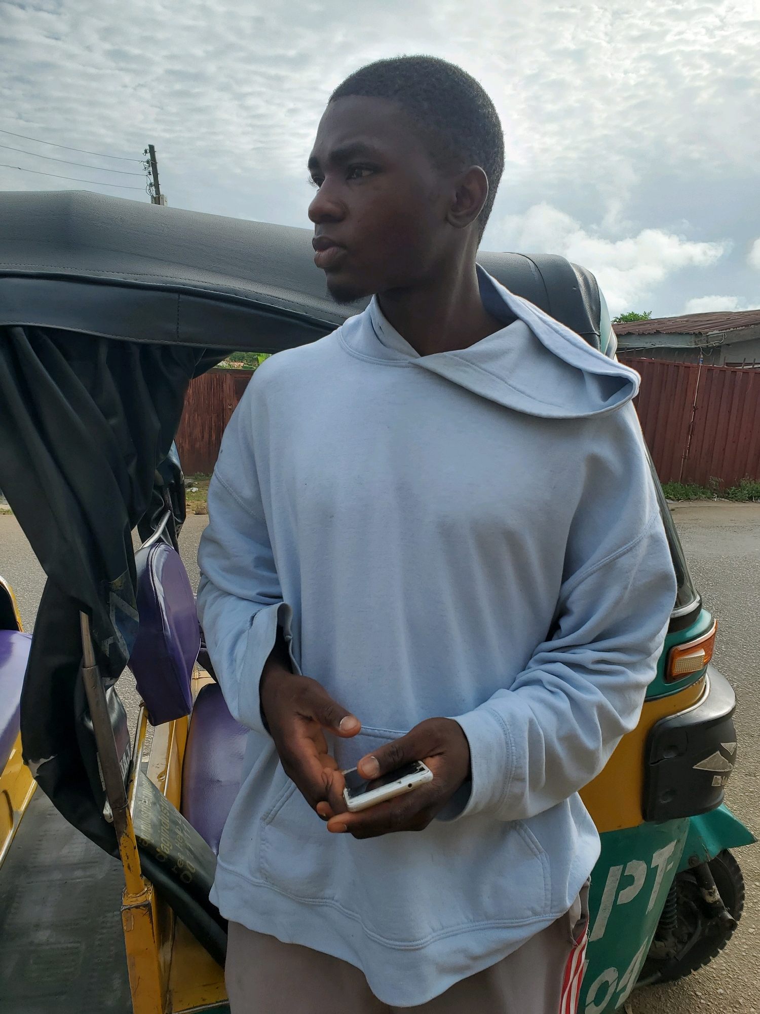 A young Black male is standing next to a three-wheeled auto-rickshaw, possibly in Jos, Nigeria. He is wearing a light blue hooded sweatshirt and light-colored pants. His left hand is holding a white smartphone. The auto-rickshaw has a black canopy and purple seats. The background shows a cloudy sky, a fence, and some buildings. The overall impression is a candid street-style shot.