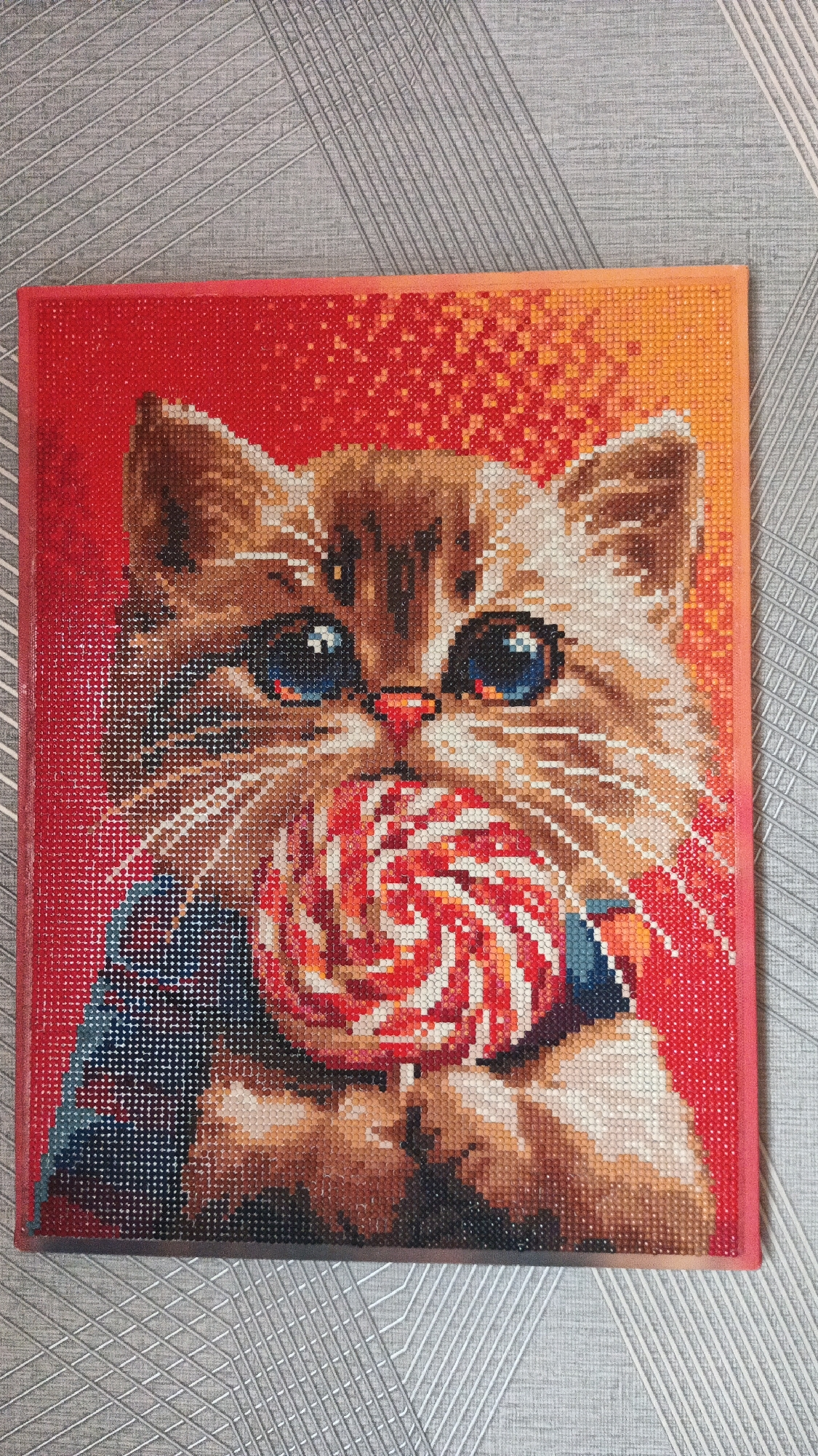 A diamond painting of a kitten holding a lollipop is displayed on a grey, textured surface. The kitten has blue eyes, brown and white fur, and is wearing a blue and dark red garment. The background of the painting transitions from red to orange. The diamond painting is framed in red and appears to be approximately 12x16 inches in size. The image is captured from a slightly elevated angle, showing the top edge of the painting. There is no discernible connection to Kryvyi Rih, Ukraine, or any specific city.