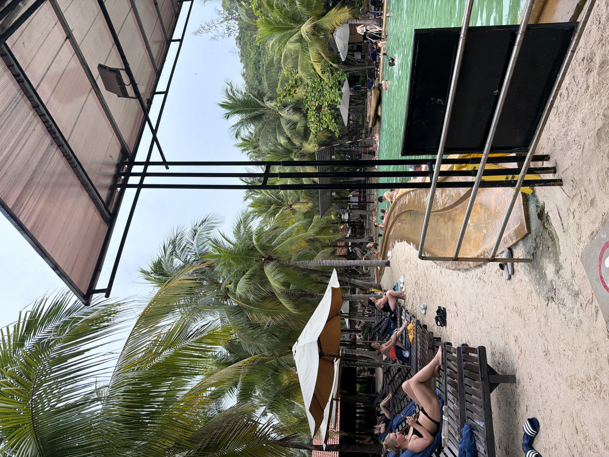 The image depicts a lively scene at a resort pool in Nha Trang, Vietnam, likely a water park or spa. Numerous palm trees surround a large, turquoise pool where many people are swimming and relaxing. The pool area is sandy, with several lounge chairs and umbrellas scattered throughout. In the foreground, people are lounging on chairs, some reading or looking at their phones, while others are walking. The overall atmosphere appears to be one of leisure and recreation, with a bright, tropical feel.