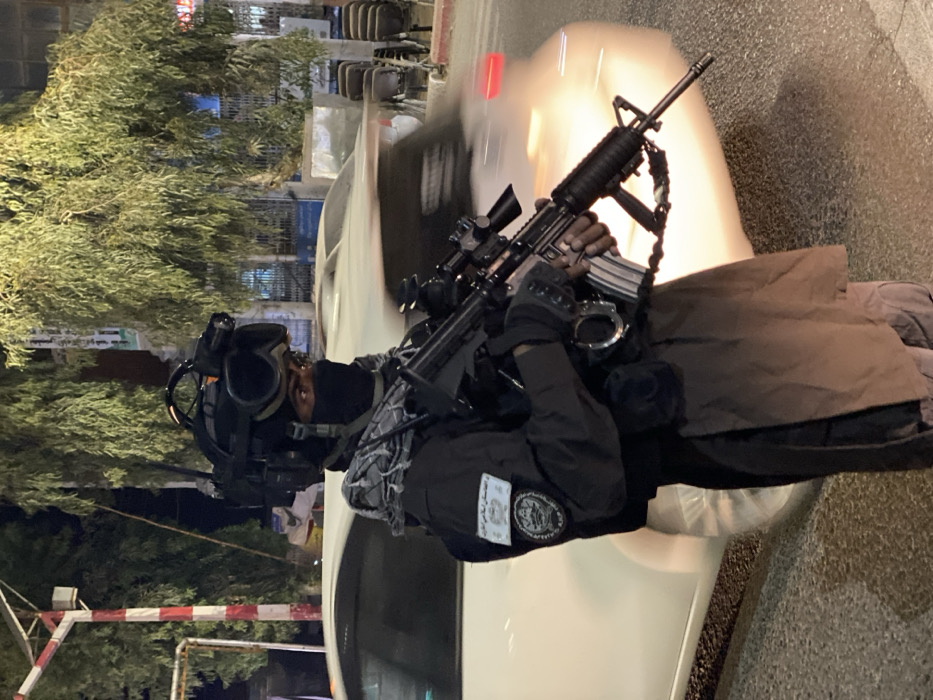 A male individual in tactical gear stands on a paved street at night in Kandahar, Afghanistan. He wears a black ballistic helmet with a mounted tactical light and communication equipment, dark goggles, and a black balaclava that covers his face, exposing only his eyes. A grey and black patterned scarf is wrapped around his neck. His dark-colored uniform includes a shirt with a rectangular patch displaying Arabic script and an emblem on the left sleeve, with a circular patch directly below it. He is armed with an M16/M4-style assault rifle, equipped with a scope and loaded with a magazine, held in a ready position. Metal handcuffs are attached to his vest. In the background, a white sedan is partially visible, blurred by motion, on the roadway. The street is lined with trees and illuminated by multiple artificial light sources. Stacked chairs are present on the right side of the roadway.