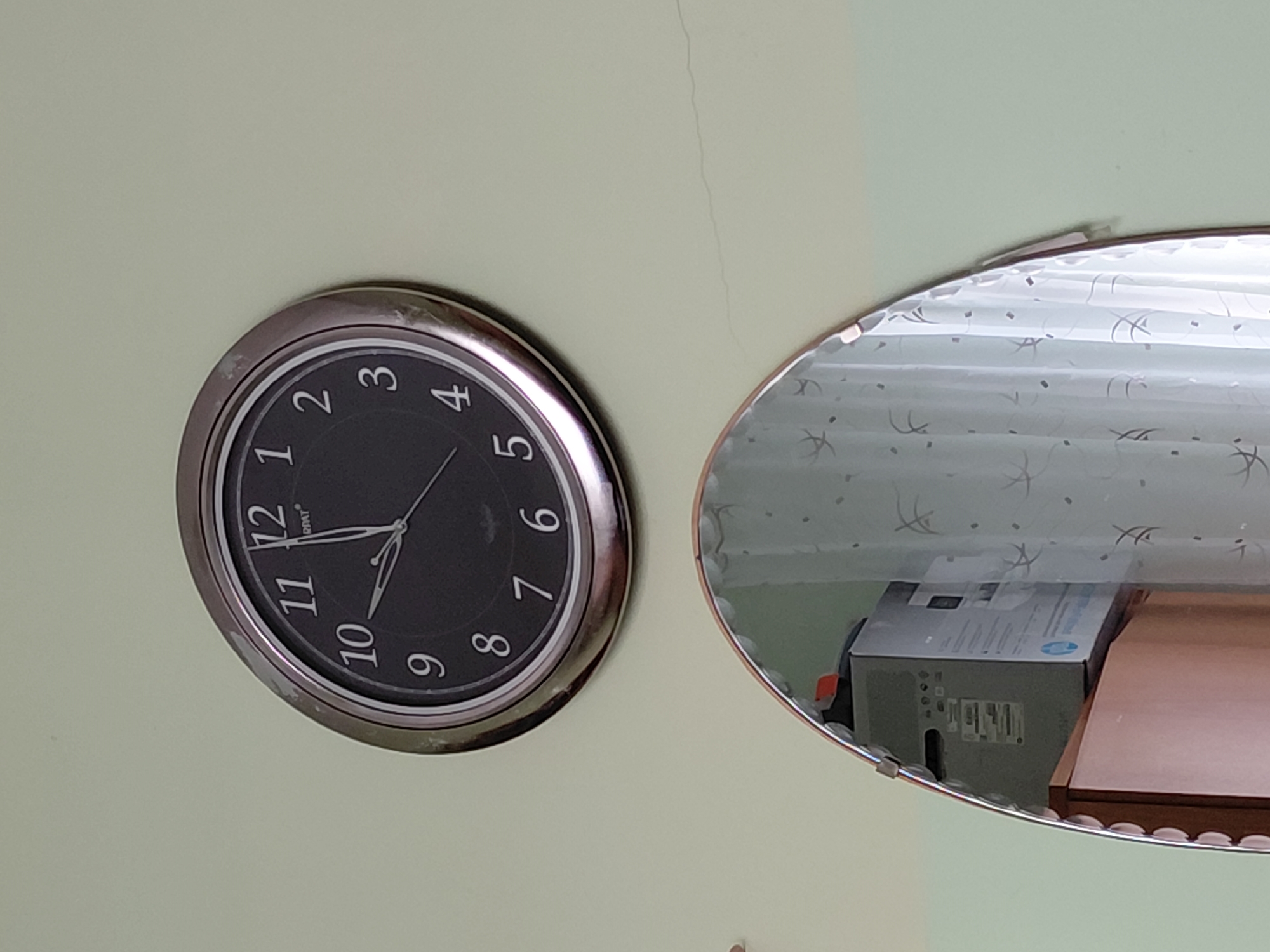 The image presents an indoor scene featuring a wall-mounted analog clock and an adjacent, irregularly shaped mirror. The wall is painted a light, muted green or pale yellow hue, with a subtle vertical crack visible above and to the right of the clock.
The round clock has a silver-colored metallic frame, showing some signs of wear. Its black face features clear white Arabic numerals from 1 to 12. The time displayed by the white hour and minute hands is approximately 9:45, with the hour hand positioned between 9 and 10, and the minute hand directly on the 9. A slender white second hand is also visible. Small white text, likely a brand name "RIFAT", is printed above the 12 numeral.
To the right of the clock, the mirror, with a thin metallic trim, reflects a portion of the room. The reflection clearly shows white or light-colored curtains adorned with a repeating pattern of abstract shapes or possibly stars. Below the curtains, a grey cardboard box, possibly packaging for an electronic device, is visible. It features barcodes and some unreadable text, with a partial "HP" logo discernible on its side. A reddish-brown wooden surface, likely a table or shelf, is partially seen behind and next to the box.
No people are present, and no specific activity is taking place. The scene is static, depicting an ordinary interior setting. No explicit visual cues within the image indicate the provided location context of Nadym, Russia, nor is any specific time of day or weather discernible from the indoor environment.